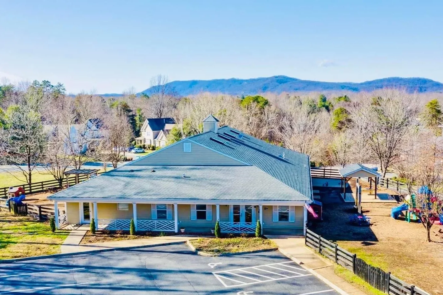 A single-story building with a metal roof, small front porch, and a parking lot in front. Surrounding the building are leafless trees, a fenced yard, and a playground area with slides. Hills or mountains are visible in the distance under a clear sky.