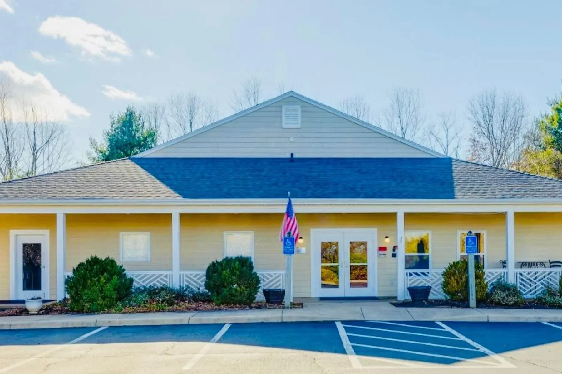 Single-story building with a beige exterior, white porch railing, and a dark gray roof, with parked spaces in front, a flagpole with an American flag, and two handicap parking signs.