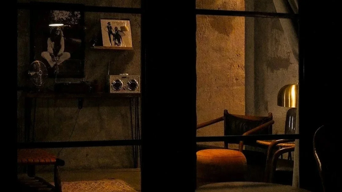 A dimly lit room viewed through a window with black framing, showing a vintage style interior with chairs, a floor lamp, and wall decorations.