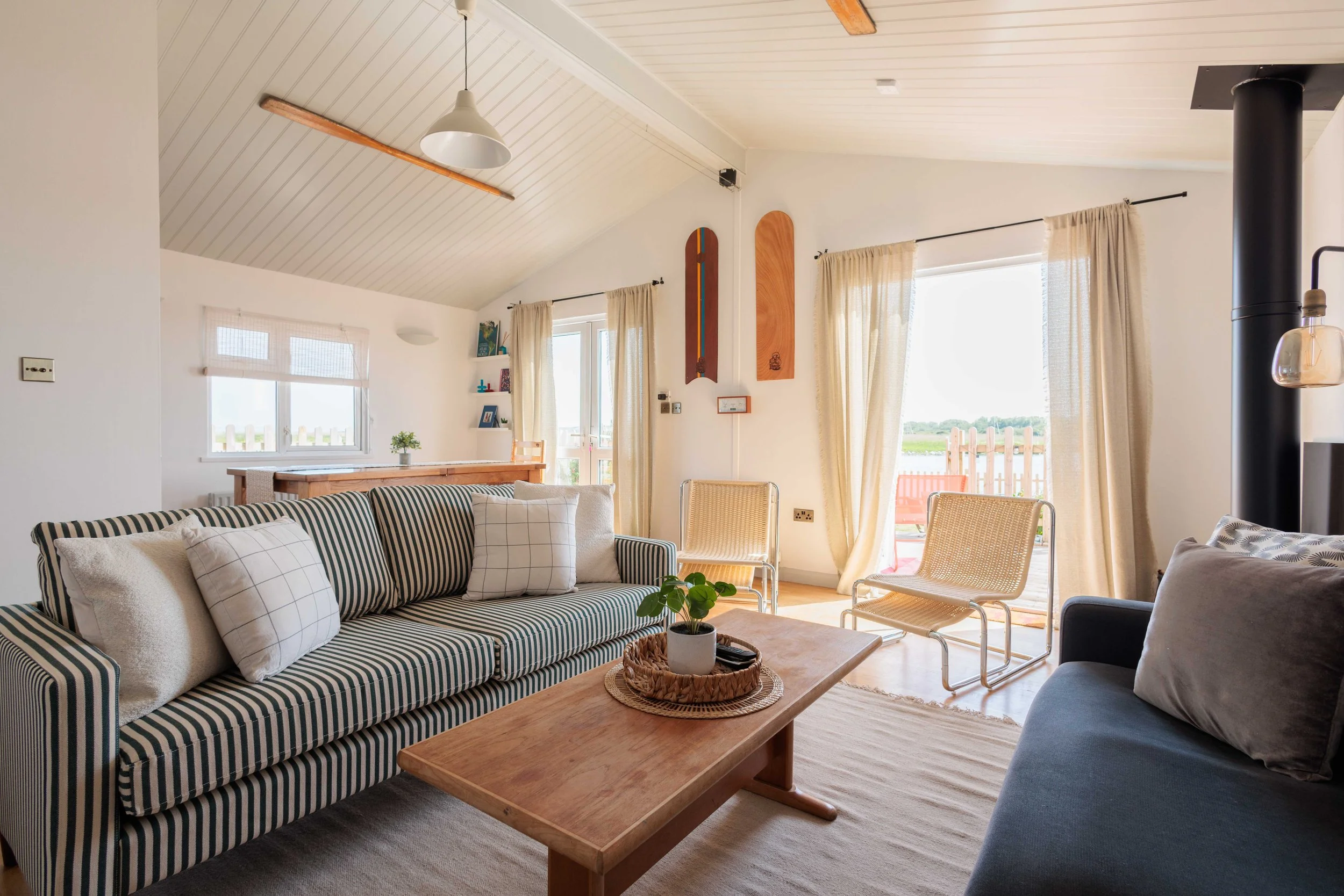 Living room with a wood-burning stove, gray sofa with throw blankets and pillows, wooden coffee table, white armchair with striped pillow, wall-mounted TV, painting of sailboats, cacti, and wall sconces providing warm lighting.