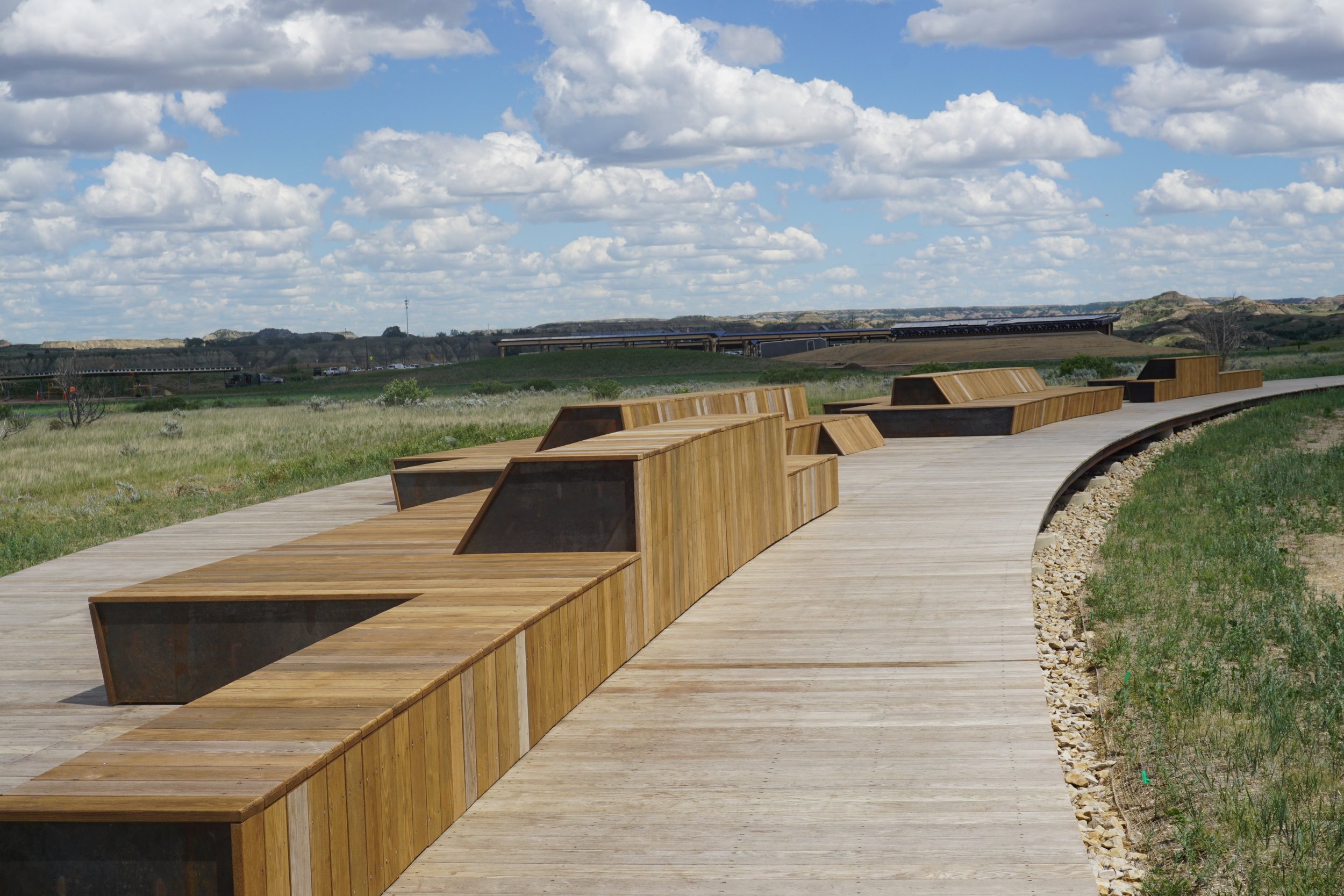 Wooden benches at the Theodore Roosevelt Presidential Library follow a curved wooden pathway in a grassy park with cloudy blue sky and distant hills.