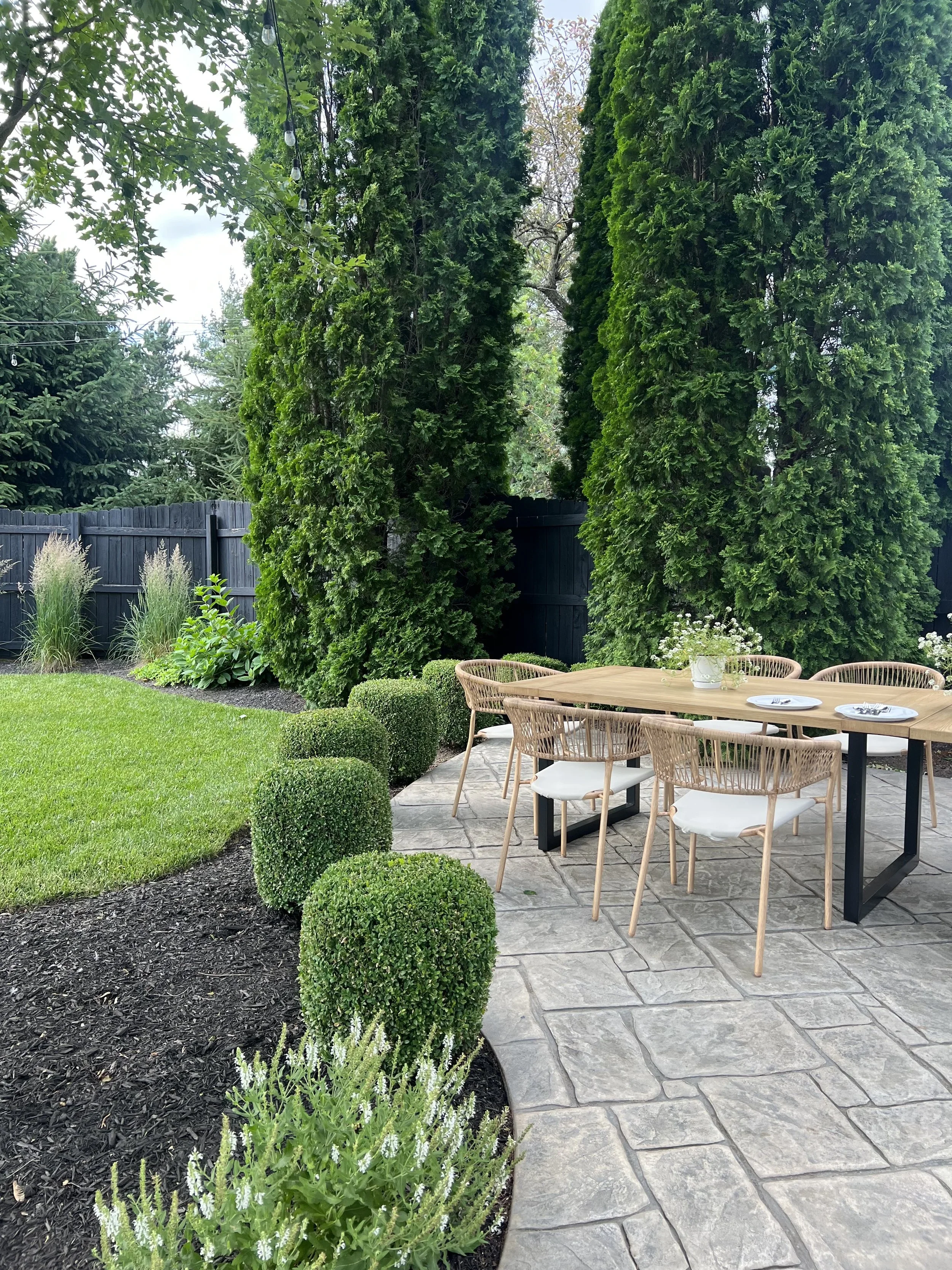 Modern backyard patio with stamped concrete, outdoor dining table and wicker chairs, manicured boxwood shrubs, arborvitae privacy trees, lush green lawn, and black wood fence landscaping design.