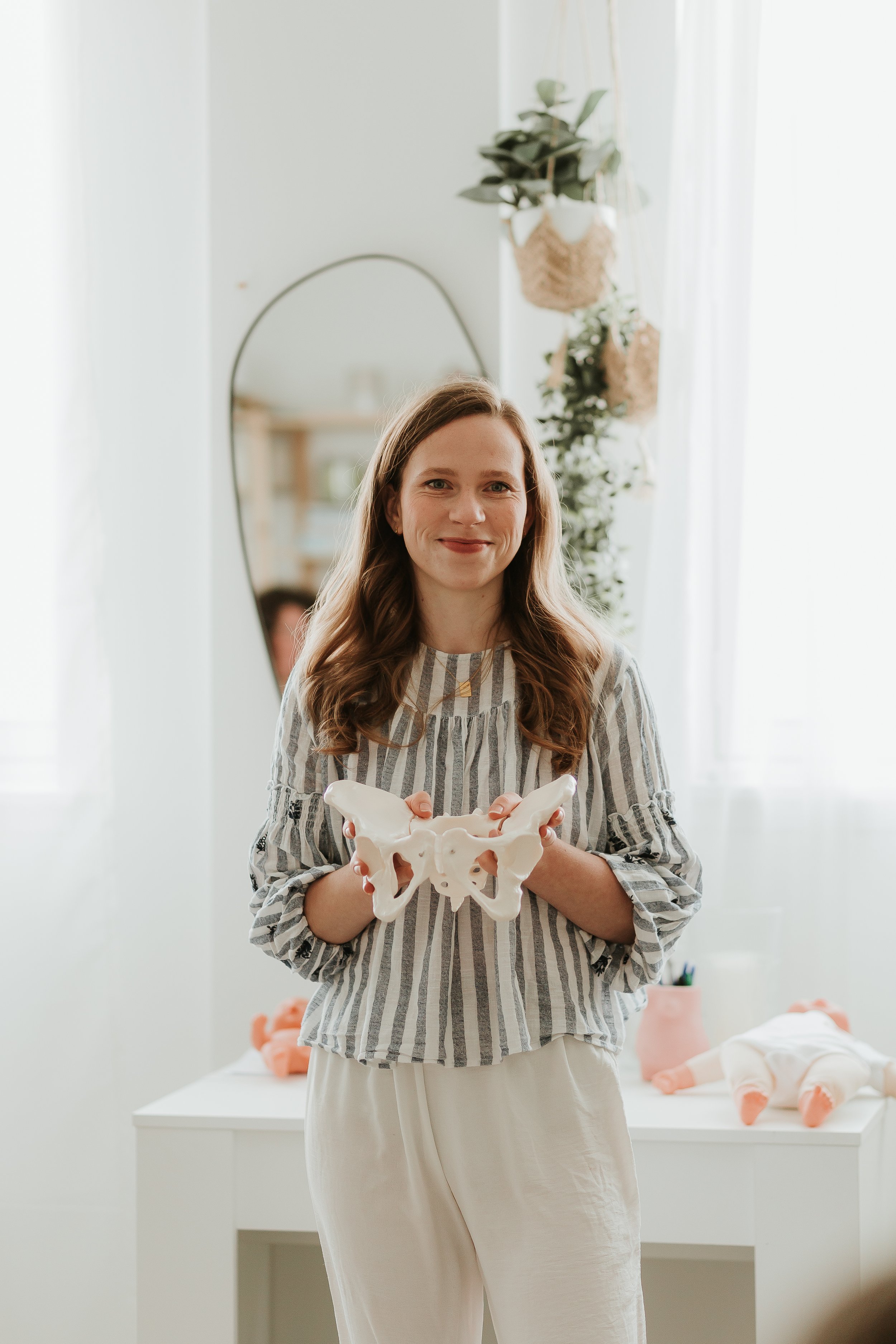 Aurelija holding a pelvis model in a room with natural light, a mirror, and plants, some dolls on a table in the background.