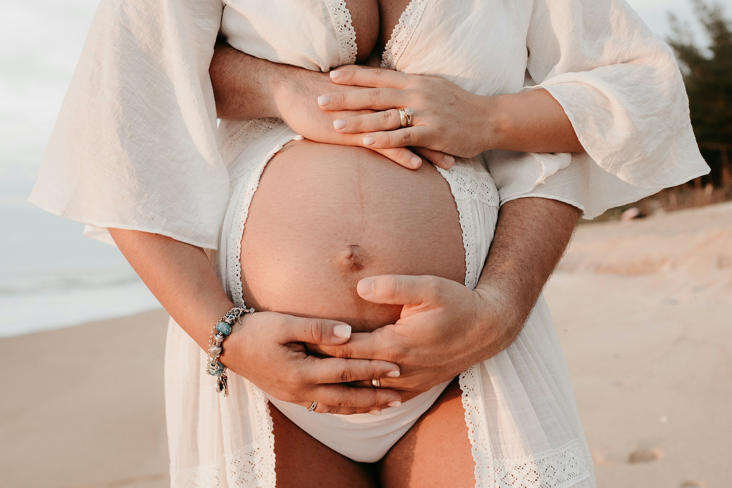 Close-up of a pregnant woman and her partner holding her belly, standing on a beach at sunset.