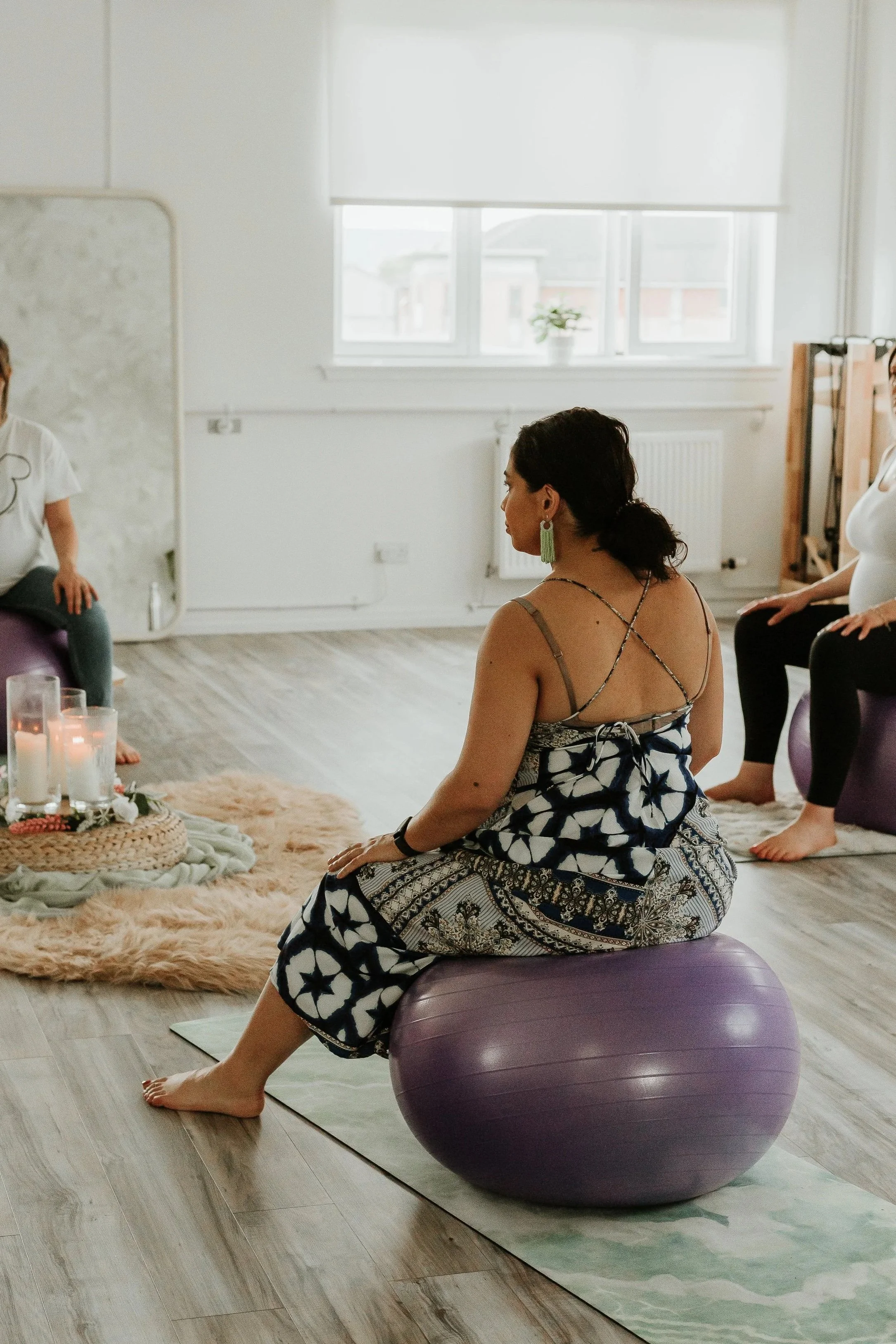 A pregnant woman sitting on a purple exercise ball in a bright room with large windows, plants, and a soft rug, participating in a pregnancy yoga or meditation session.