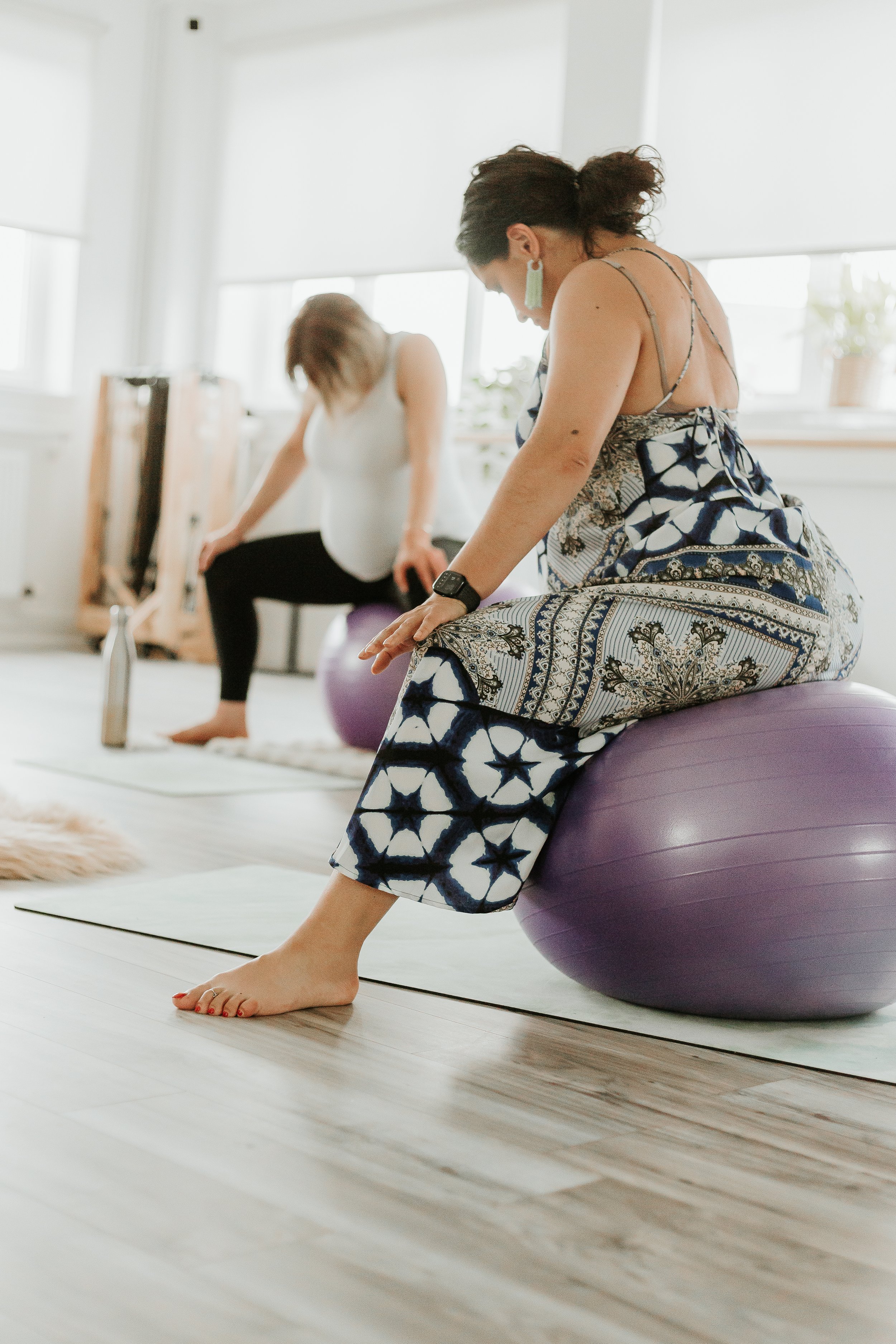 Two women participating in a birth preparation workshop doing a pregnancy yoga class, sitting on exercise balls in a bright room with large windows, wooden floors, and natural light.
