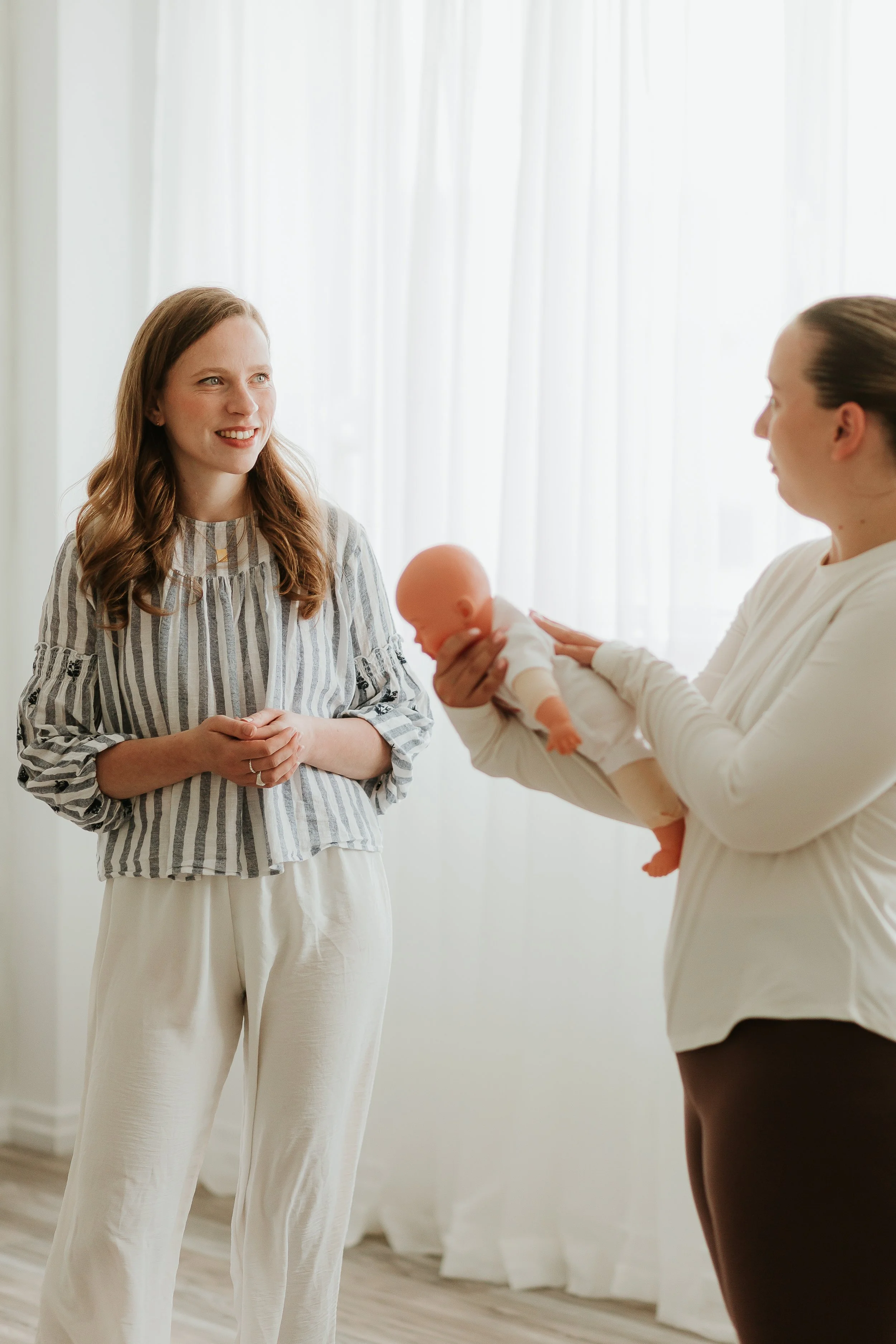 Two women, one holding a baby doll, standing in a bright room with white curtains. The woman on the left is smiling and looking at the other woman, who is holding the doll. They are having a conversation.