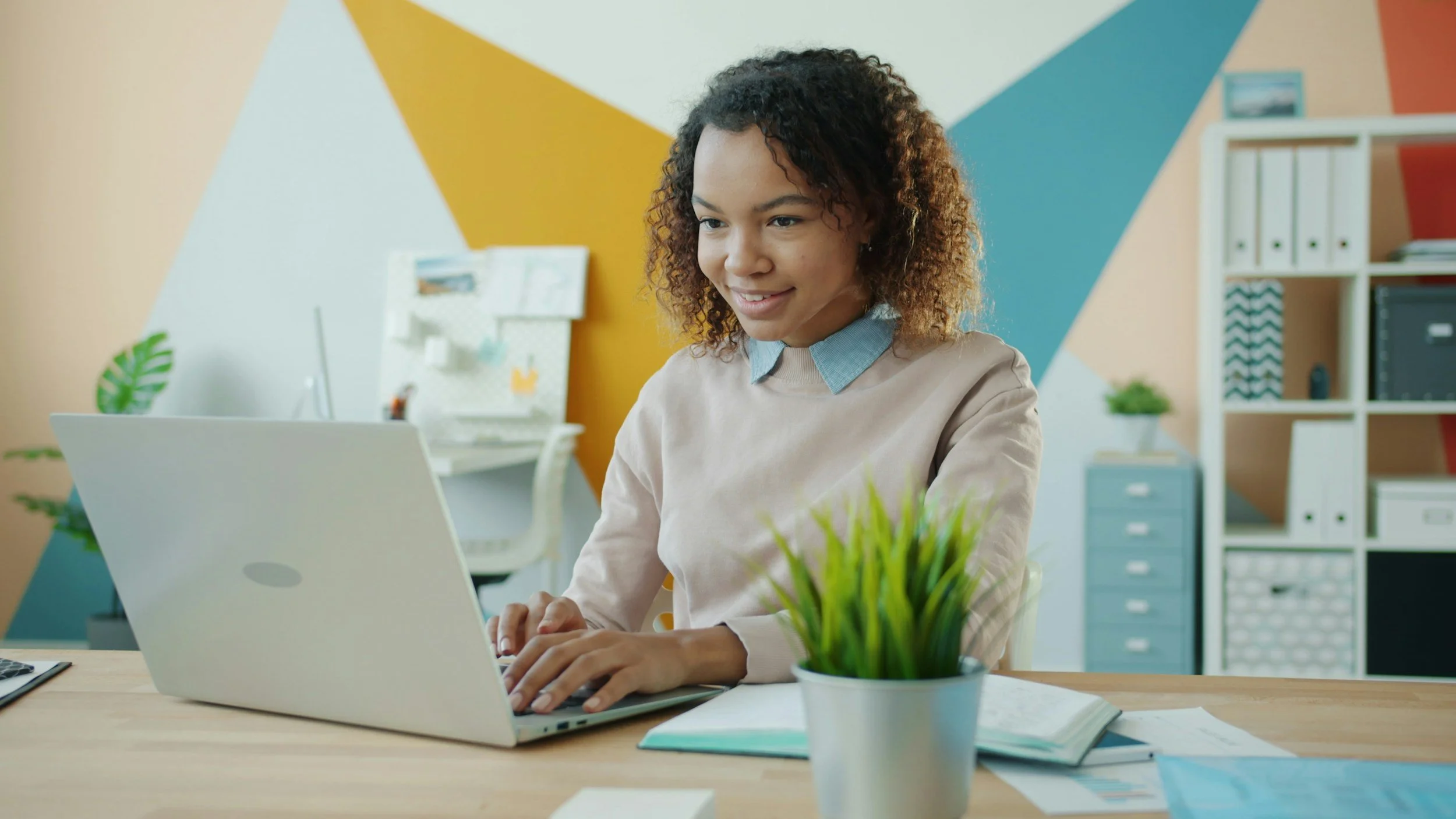 A woman with curly hair working on a laptop at her desk in a modern, colorful office. There are plants, shelves with binders, and a geometric painted wall in the background.