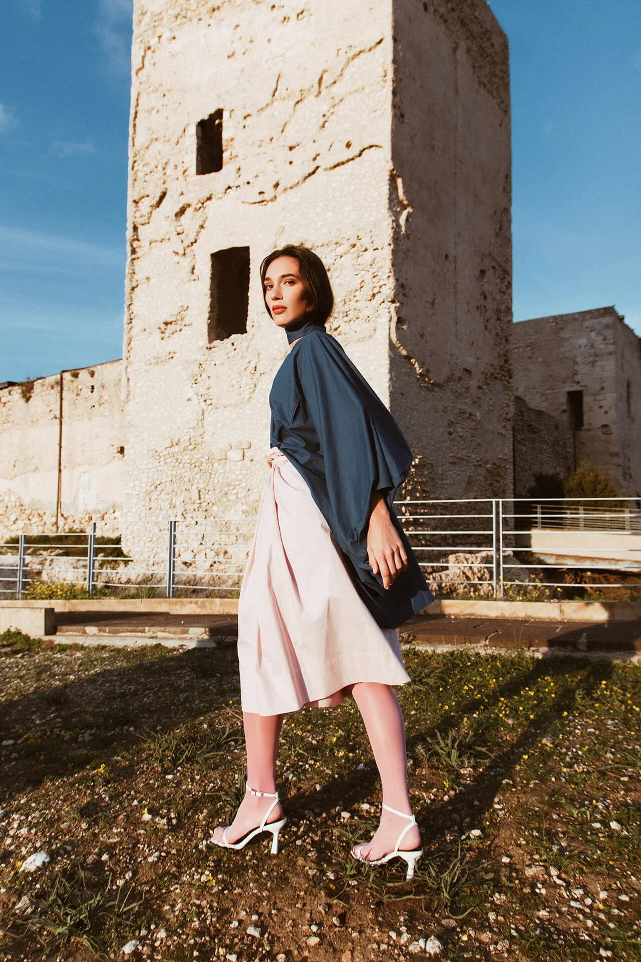 Figura intera editoriale di ragazza mora con camicia e foulard blu e gonna rosa davanti a castello Cagliari.