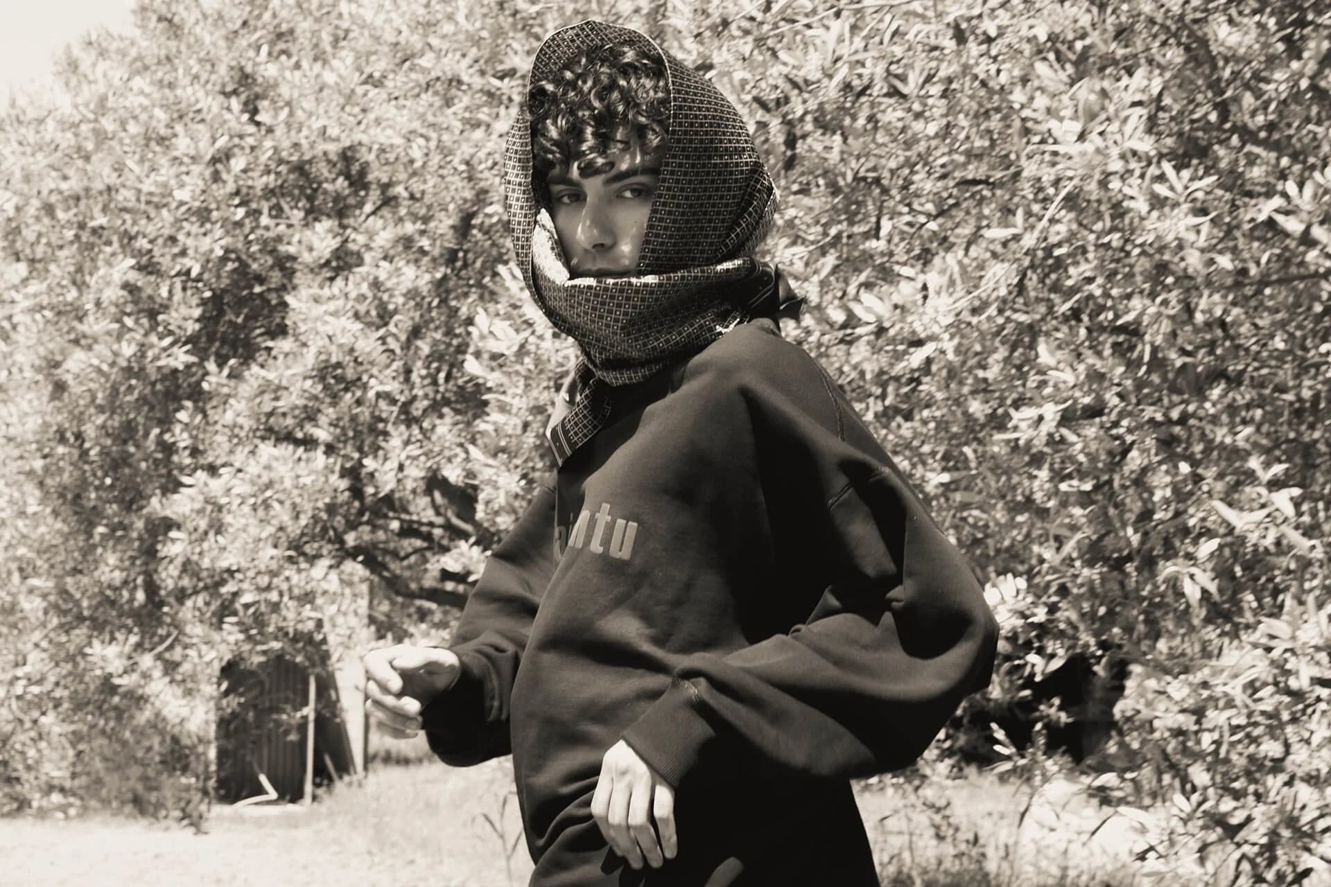 Ragazzo con cappuccio e capelli ricci in piedi in un parco, con alberi sullo sfondo, immagine in bianco e nero.