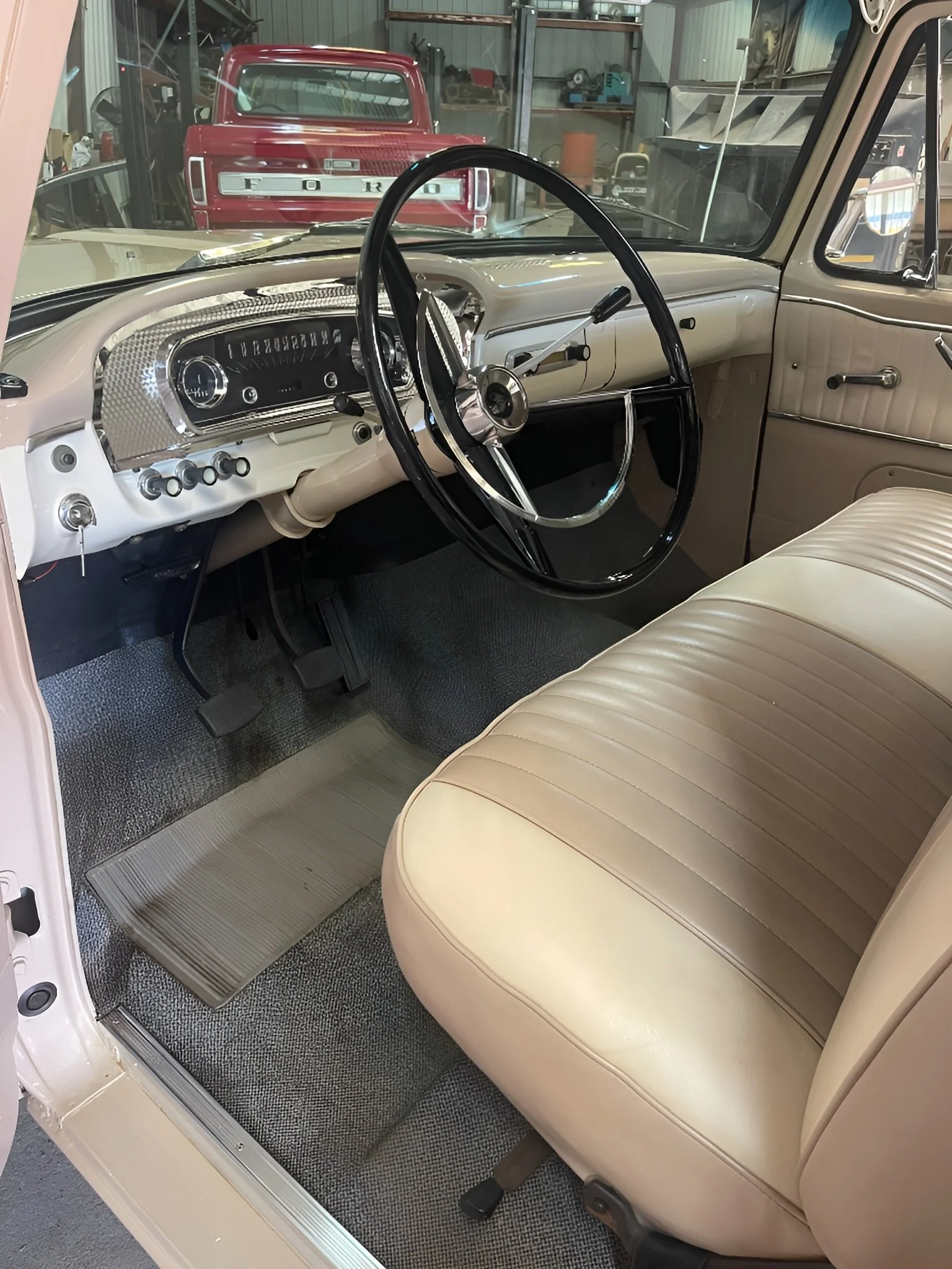Interior of a vintage car with beige seats, a black steering wheel, and a dashboard with a silver textured panel, located in a garage with a red Ford truck visible outside.