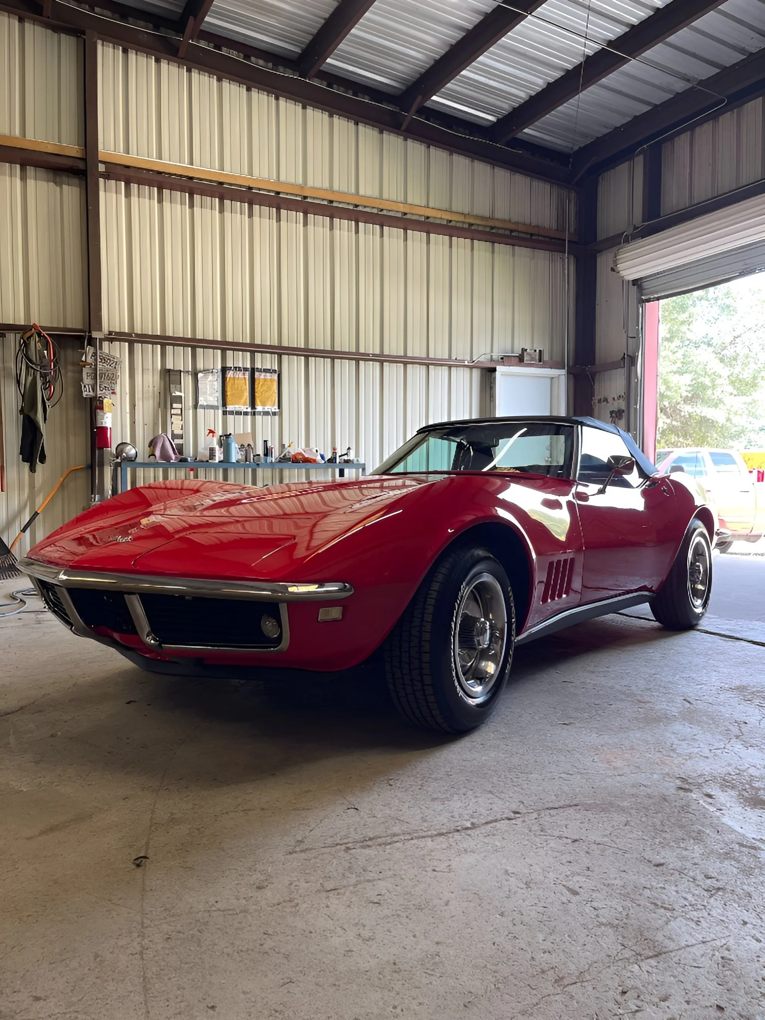 A red vintage Chevrolet Corvette sports car parked inside a large garage or warehouse with corrugated metal walls and ceiling, partially open garage door showing outside view.