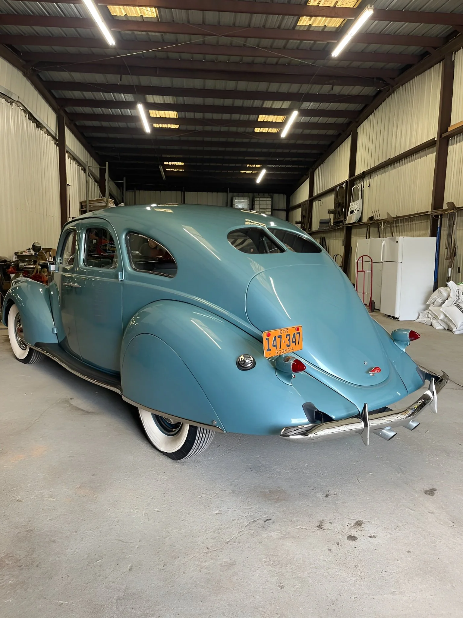 A vintage light blue car with a rounded design parked inside a large garage or warehouse, surrounded by various tools and equipment.
