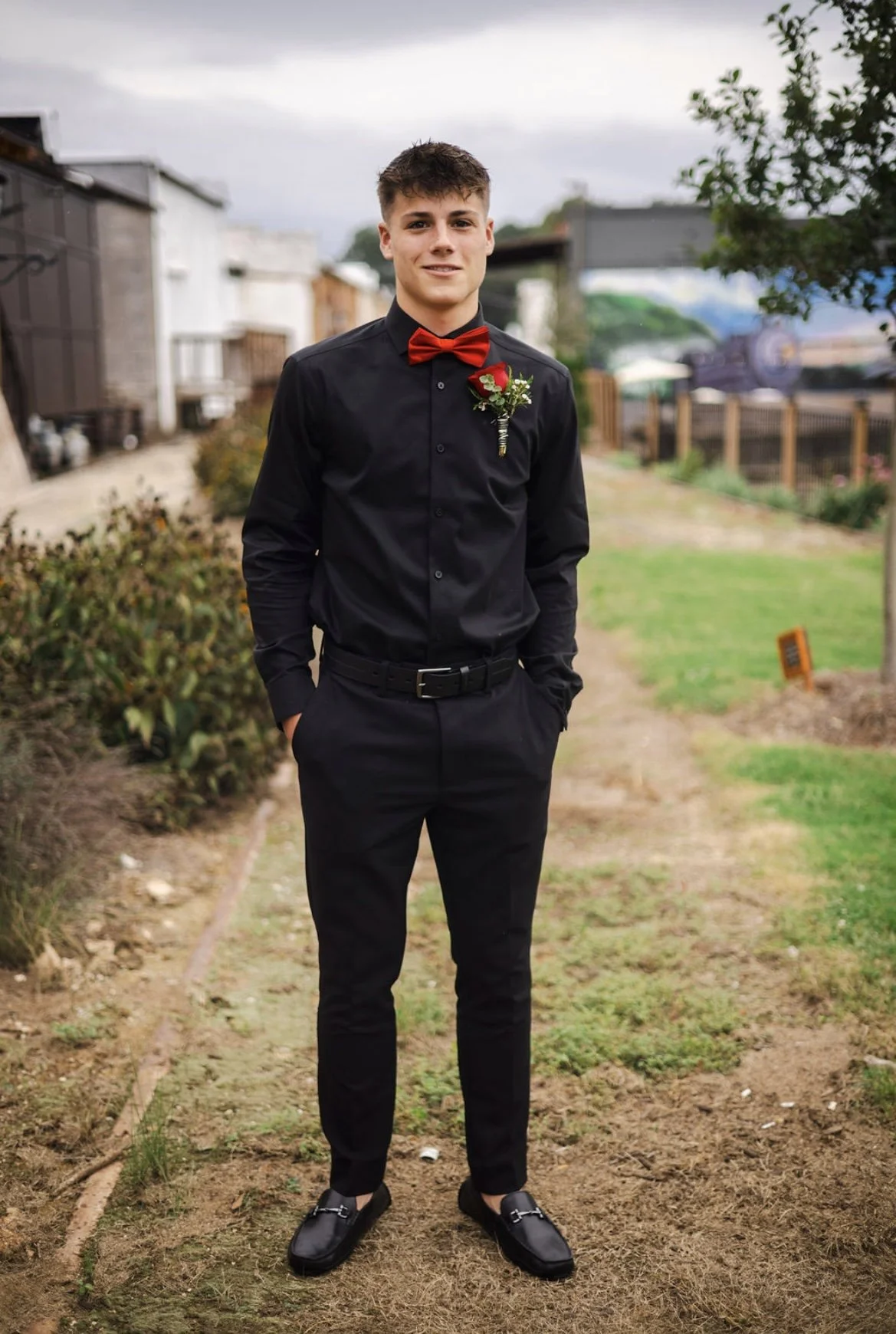 A young man wearing a black dress shirt, red bow tie, black pants, and black shoes, standing outdoors in a yard with hands in his pockets, smiling at the camera.
