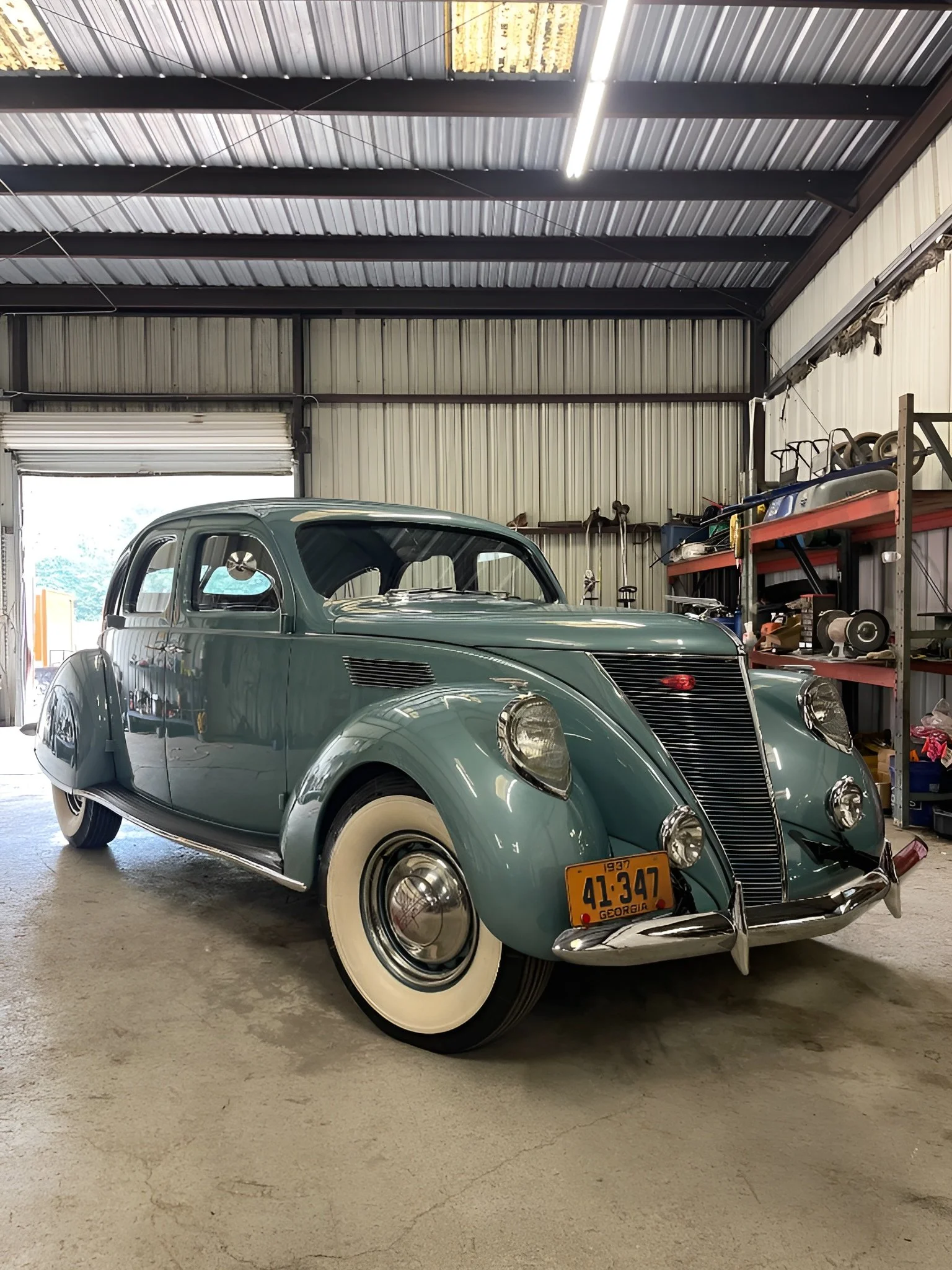 A vintage 1937 gray automobile with whitewall tires parked inside a garage or warehouse with metal shelving and equipment.