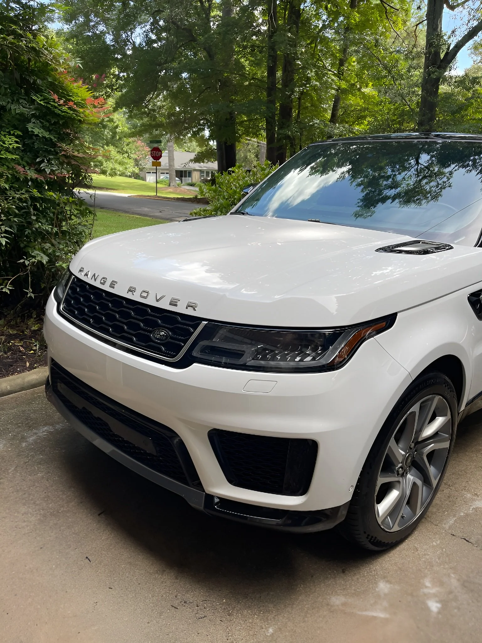 A white Range Rover parked on a driveway near green bushes and trees in a residential neighborhood, with a stop sign and houses visible in the background.