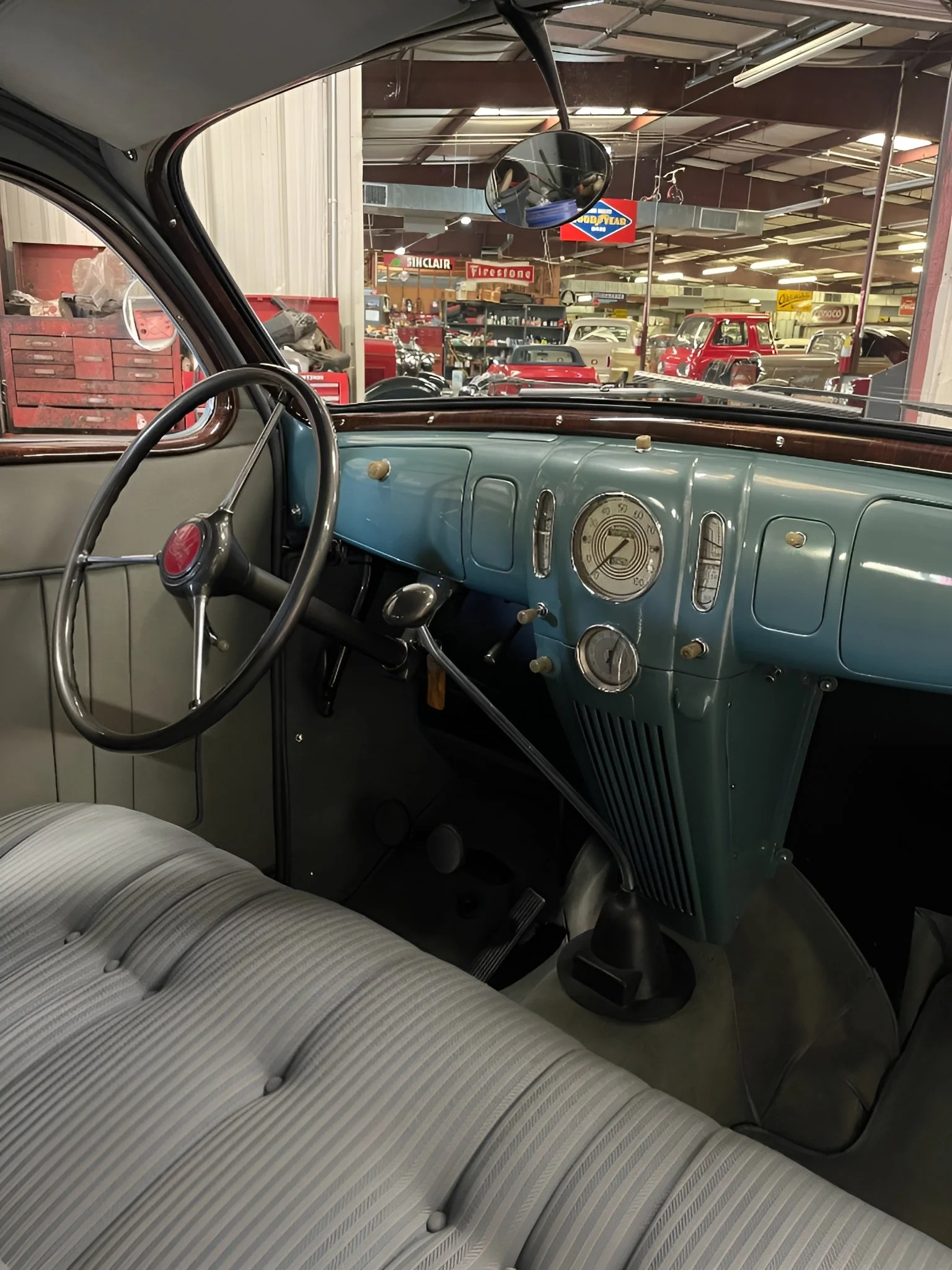 View of the interior of a vintage car, showing the dashboard, steering wheel, and bench seat in a garage or showroom with classic cars and signs in the background.