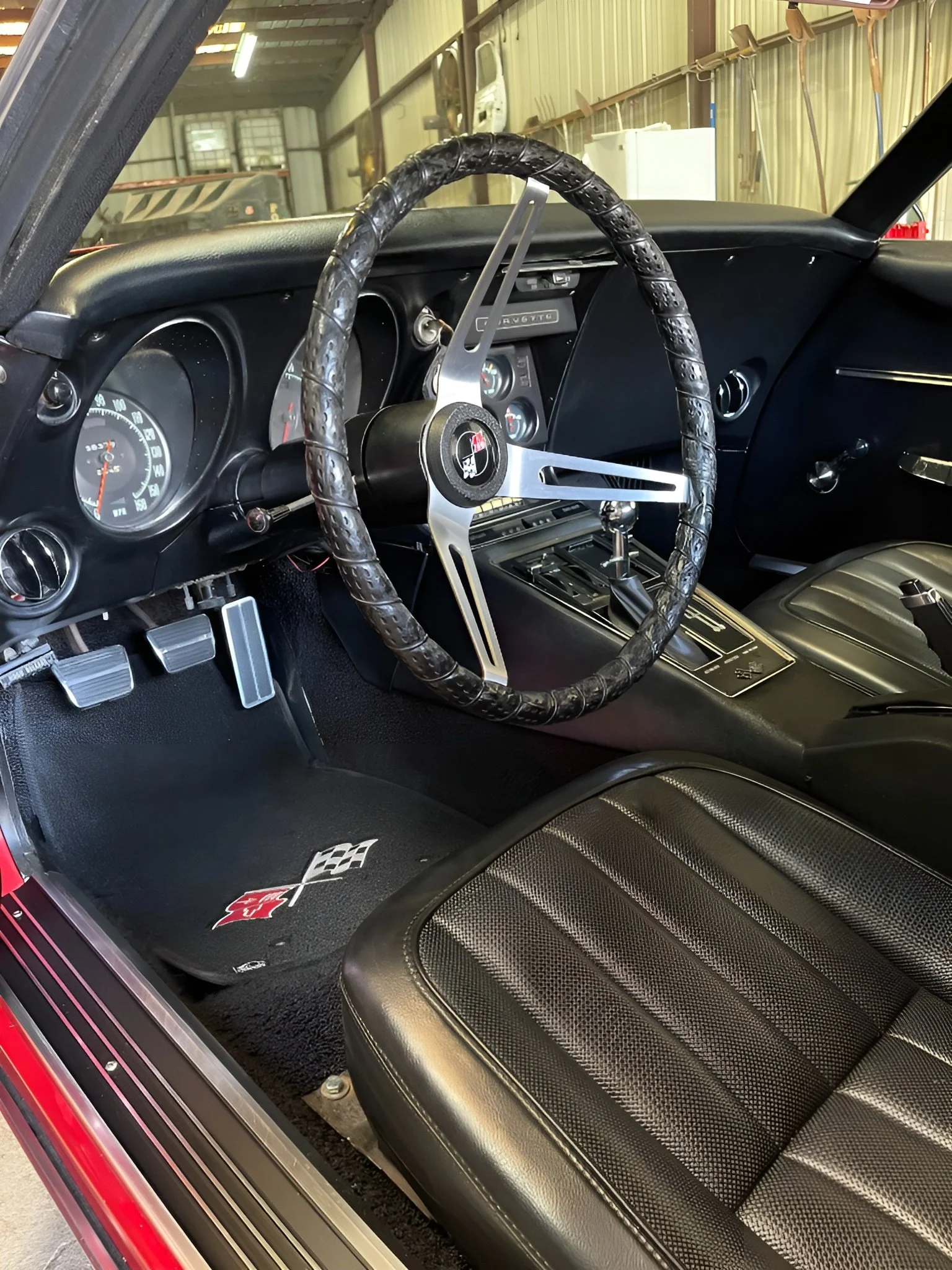 Interior of a classic Chevrolet Corvette, showing a black steering wheel with a Corvette logo, black dashboard with gauges, manual shifter, and black racing-style seat with stitching.