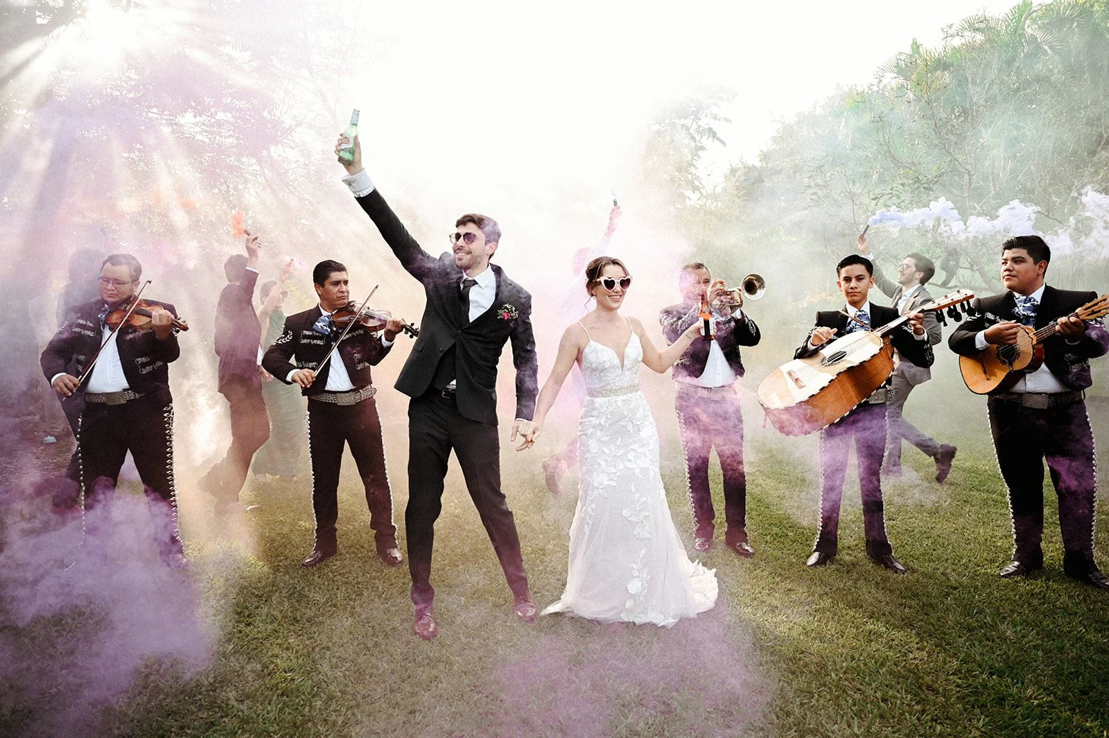 Bride and groom celebrating with a live mariachi band, colorful smoke bombs, and drinks during an energetic outdoor destination wedding in Mexico.