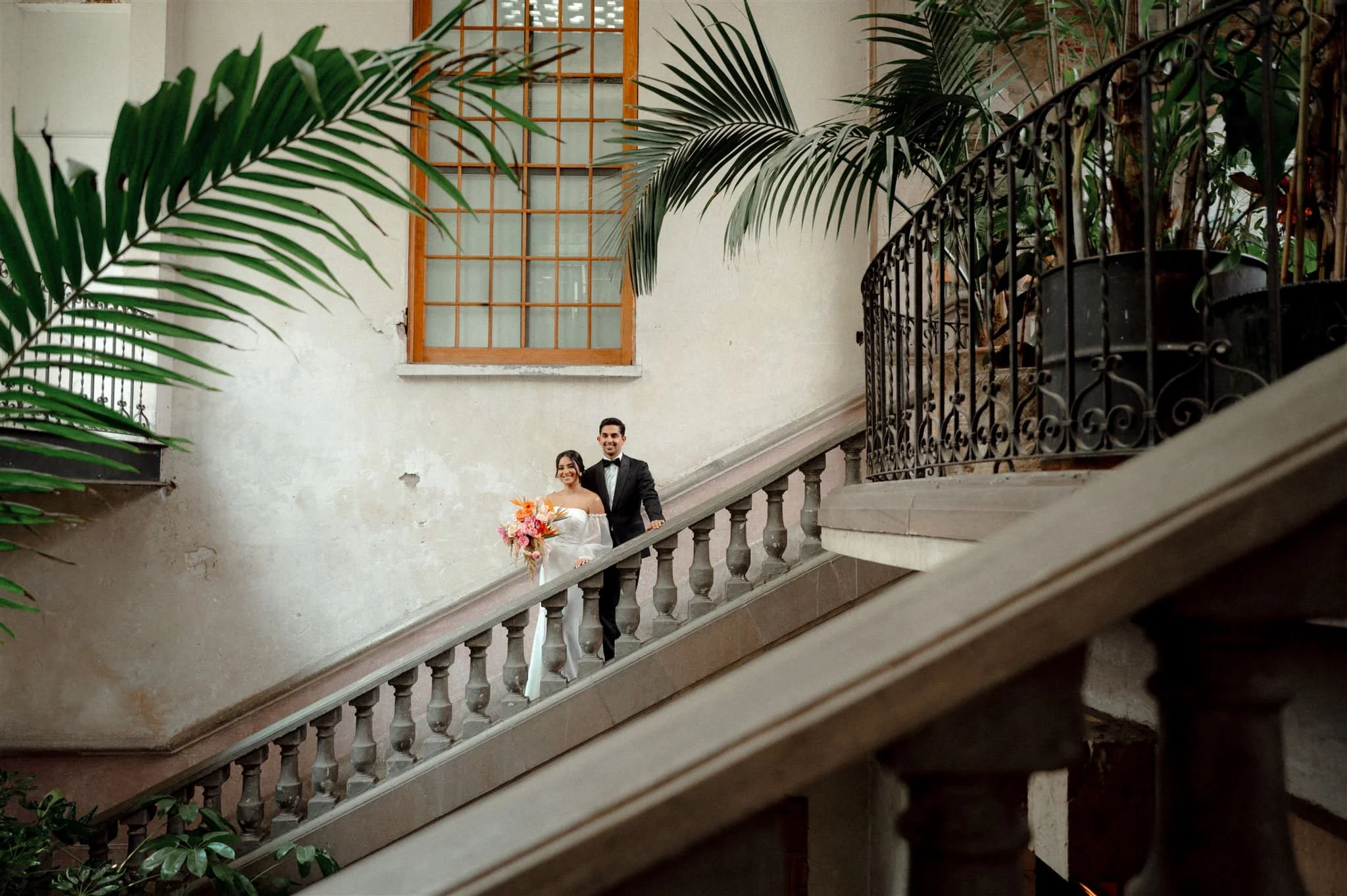 Bride and groom standing in a historic staircase at Proyectos Públicos: Prim in Mexico City, surrounded by lush greenery and elegant architecture in a modern editorial wedding scene.