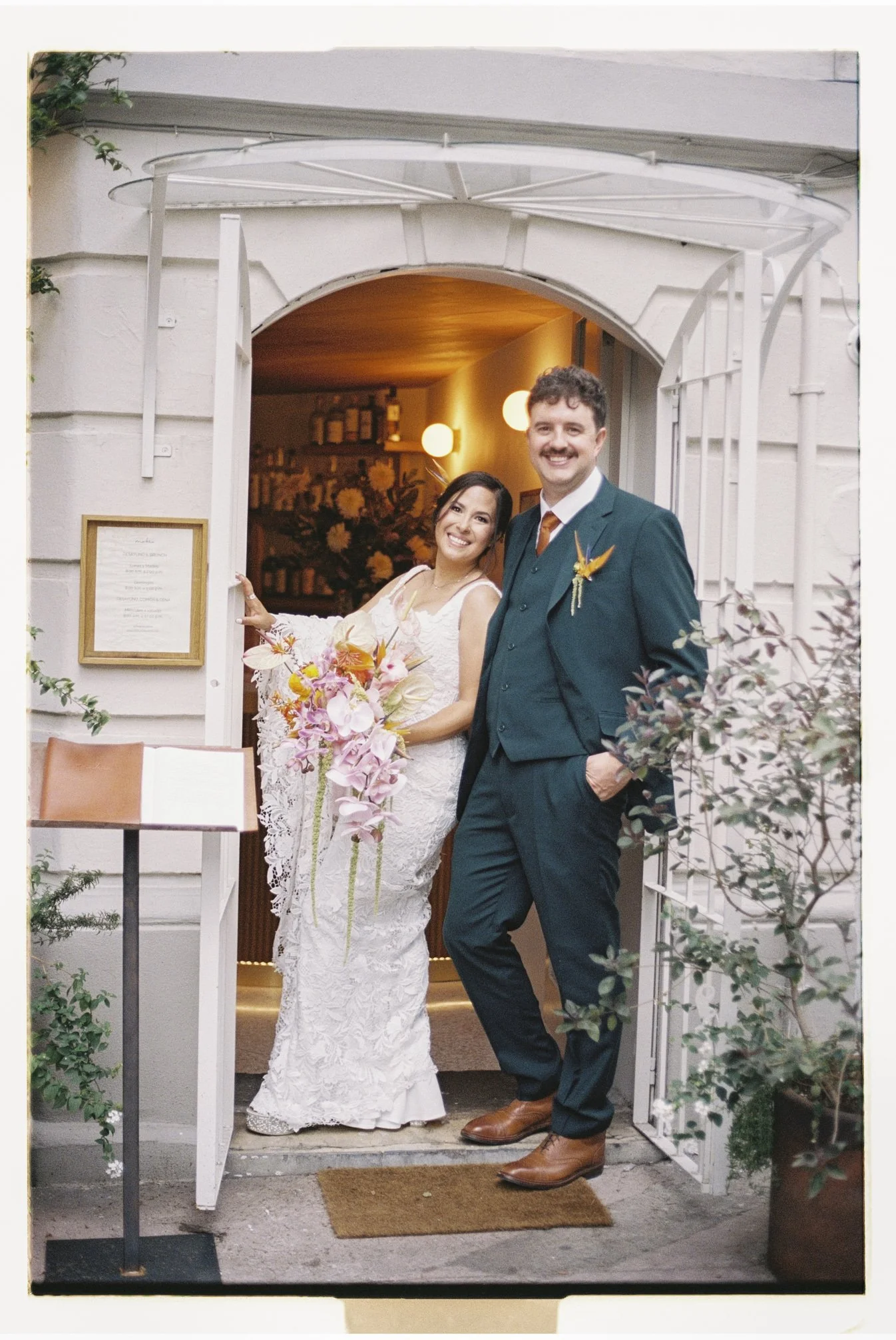 Bride and groom posing at the entrance of a boutique venue in Roma Norte, Mexico City, during an intimate elopement, featuring elegant wedding attire and colorful tropical flowers