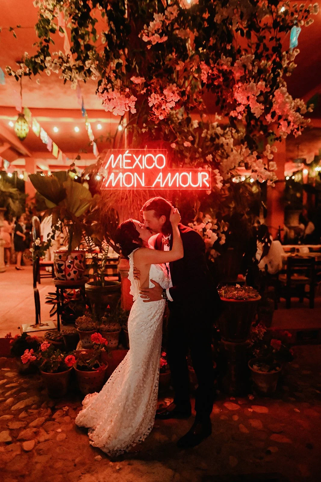 Bride and groom sharing a kiss beneath a glowing "México Mon Amour" neon sign surrounded by tropical plants and warm lighting during a destination wedding reception in Puerto Vallarta, Mexico.
