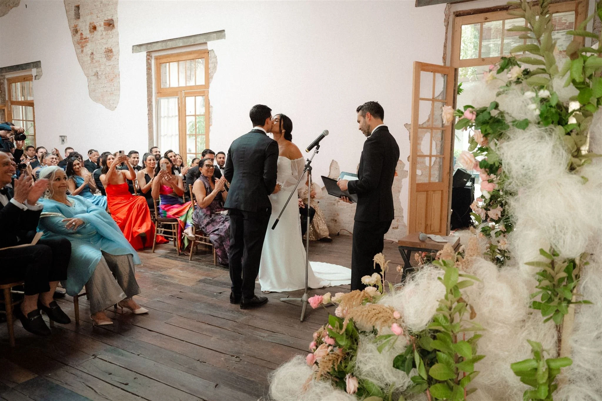 Bride and groom sharing their first kiss during the wedding ceremony at Proyectos Públicos: Prim in Mexico City, surrounded by guests, natural light, and elegant floral decor.