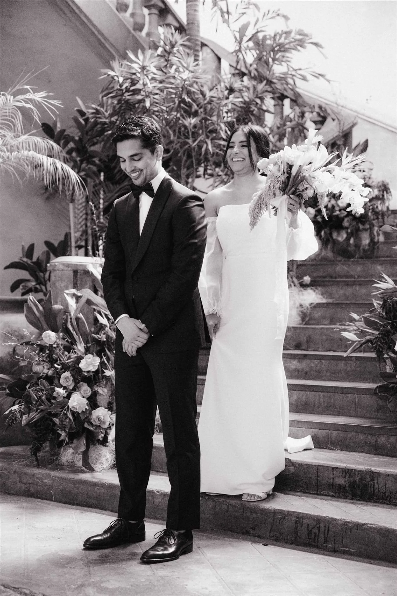 Bride surprises groom during first look on staircase at Proyectos Públicos: Prim in Mexico City, surrounded by lush greenery and floral arrangements in a romantic black and white scene.