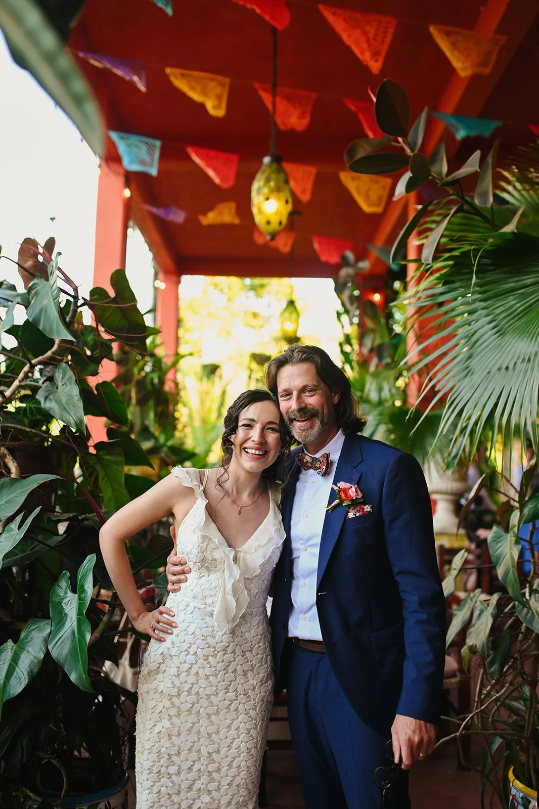 Bride and groom smiling together in a lush tropical setting at the Puerto Vallarta Botanical Garden, surrounded by colorful papel picado decorations and vibrant greenery during their destination wedding in Mexico.