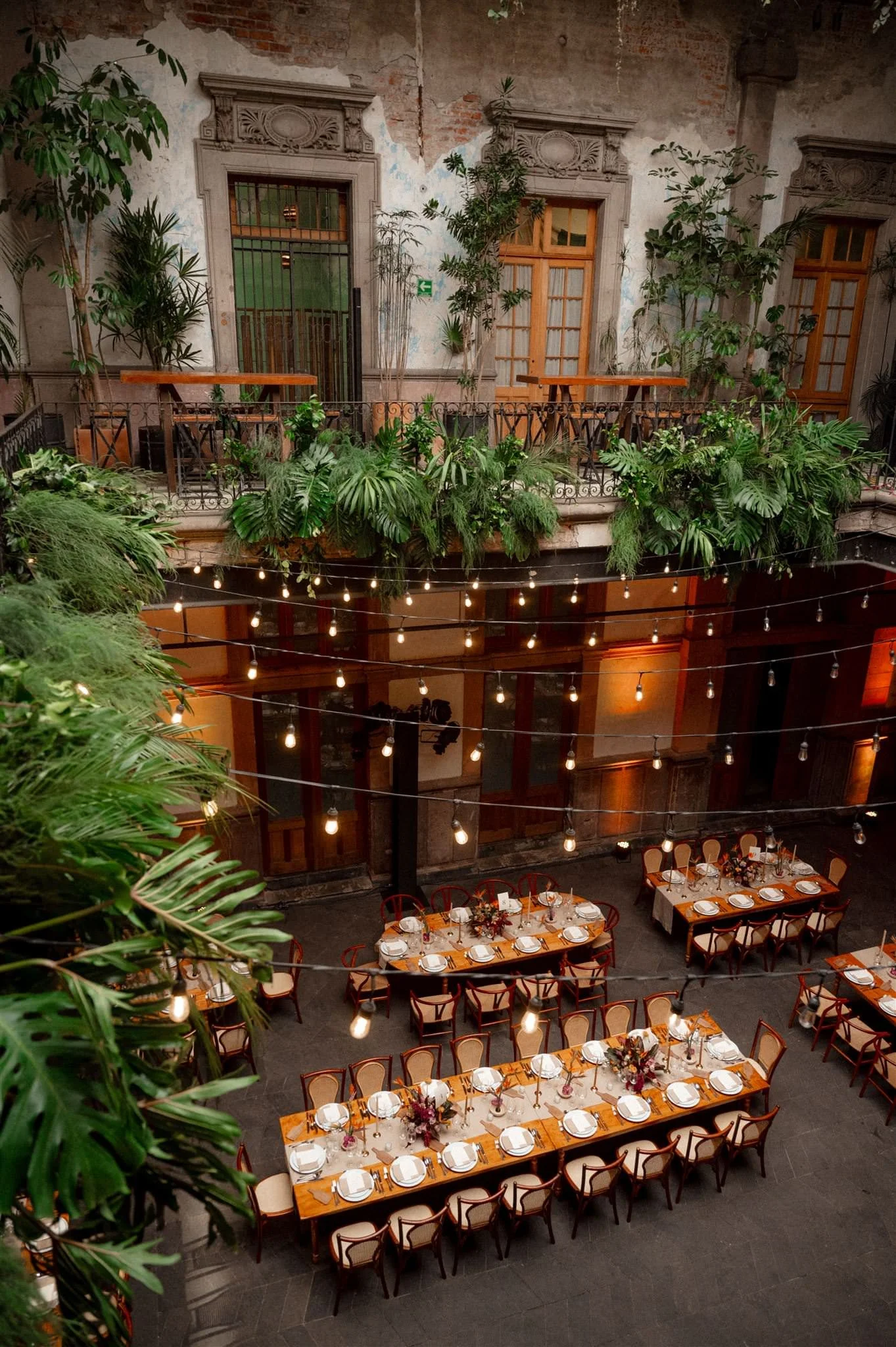 Elegant courtyard reception setup at Proyectos Públicos: Prim in Mexico City featuring long wooden tables, warm string lights, and lush greenery cascading from historic balconies for a romantic wedding atmosphere.