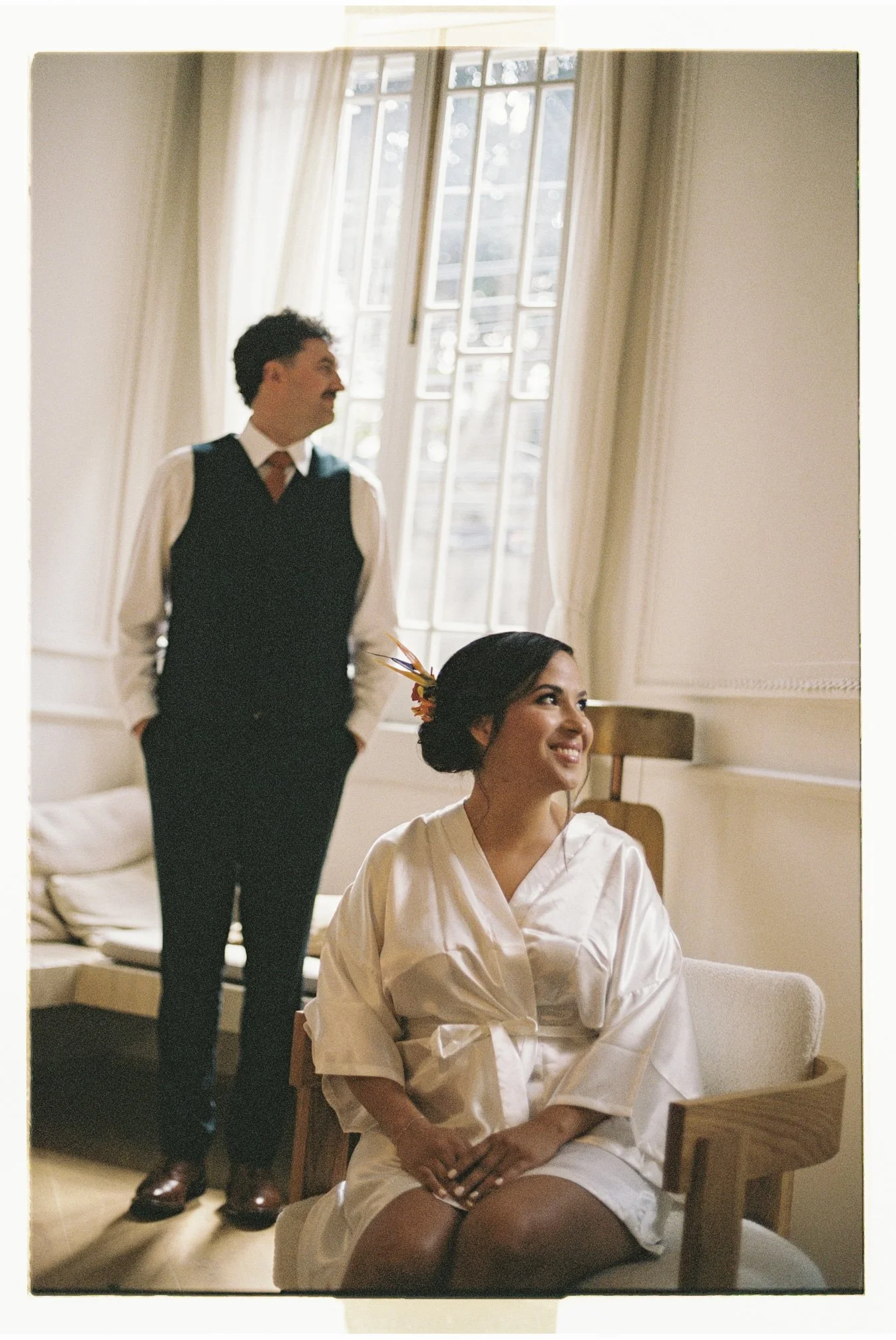 Bride sitting in a chair wearing a silk robe while getting ready for her elopement in Roma Norte, Mexico City, illuminated by soft natural window light as her partner stands nearby.