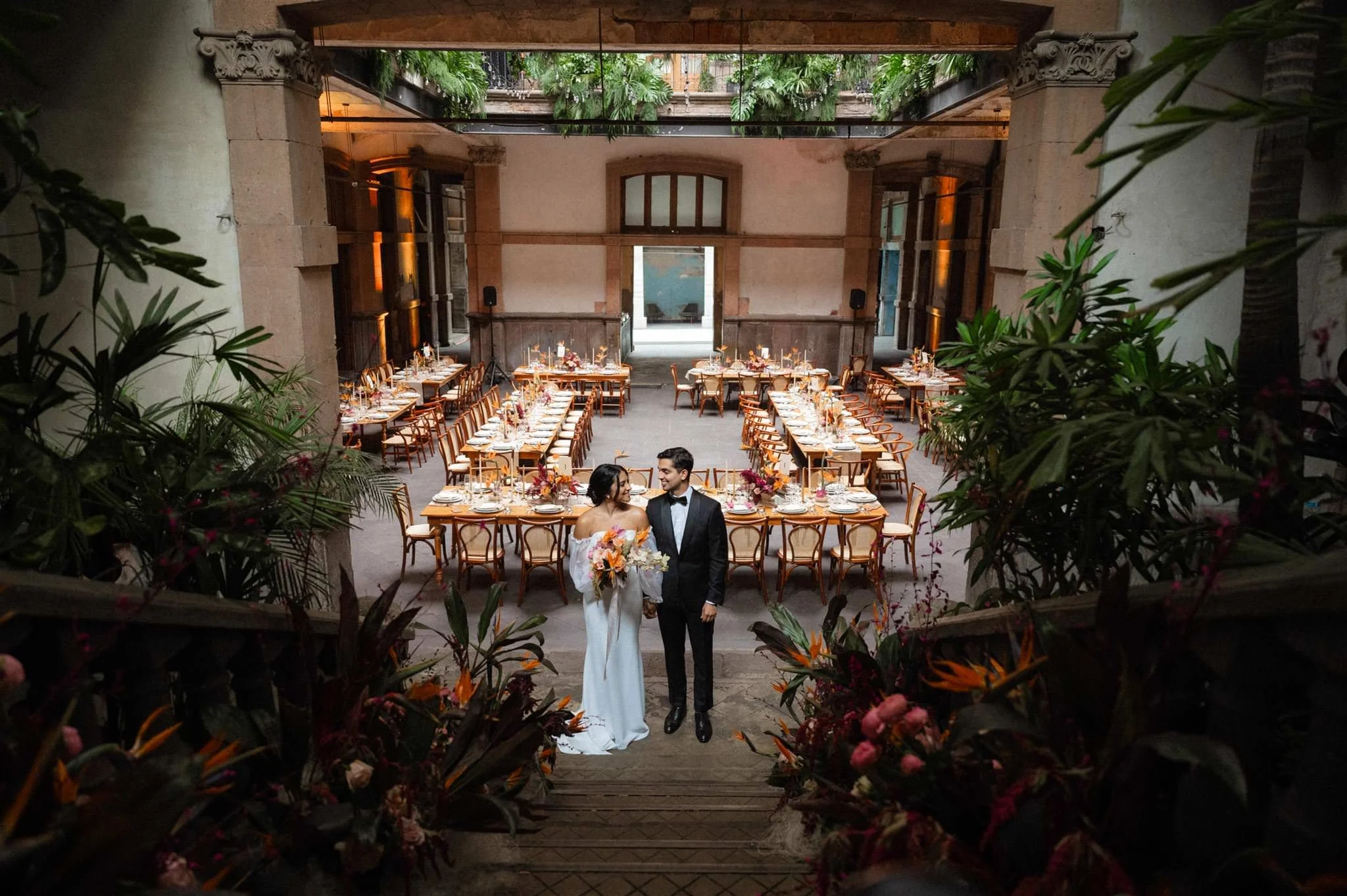 Bride and groom sharing a kiss on a historic staircase at Proyectos Públicos: Prim in Mexico City, with tropical plants and vibrant floral decor creating an intimate wedding atmosphere.