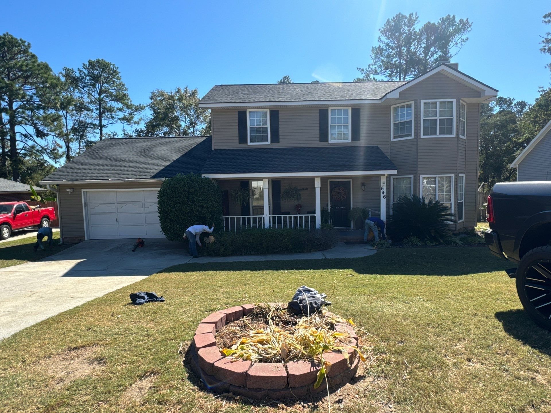 A two-story house with beige siding and white trim, with a front porch and bay window, is being landscaped by four workers. The yard has a fire pit ring made of red bricks with dried plants inside, and there are trucks parked nearby on sunny day.