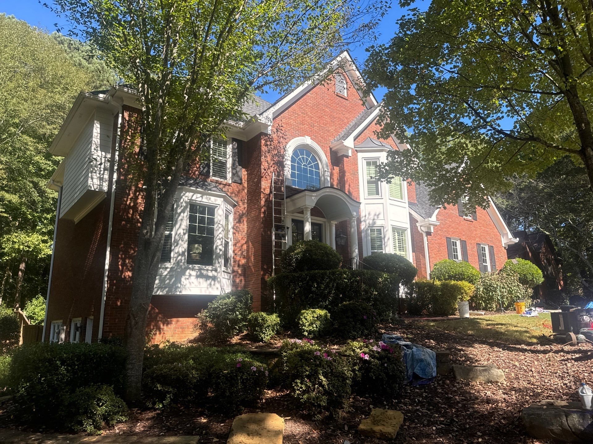 A large brick house with white trim and multiple windows, surrounded by landscaped bushes and trees on a sunny day.