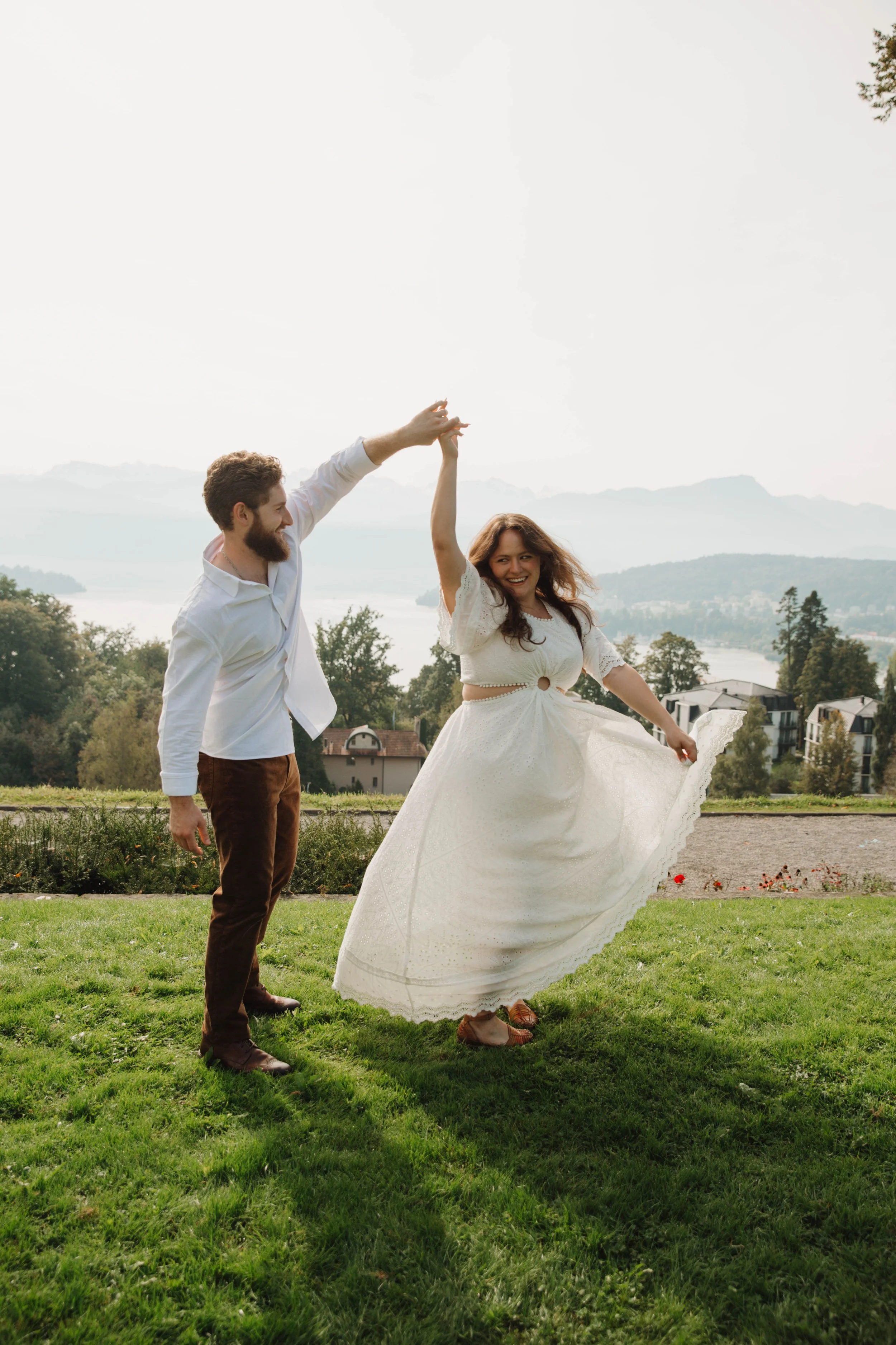 Man twirling his fiancée in a white dress at a garden overlook above Lake Lucerne
