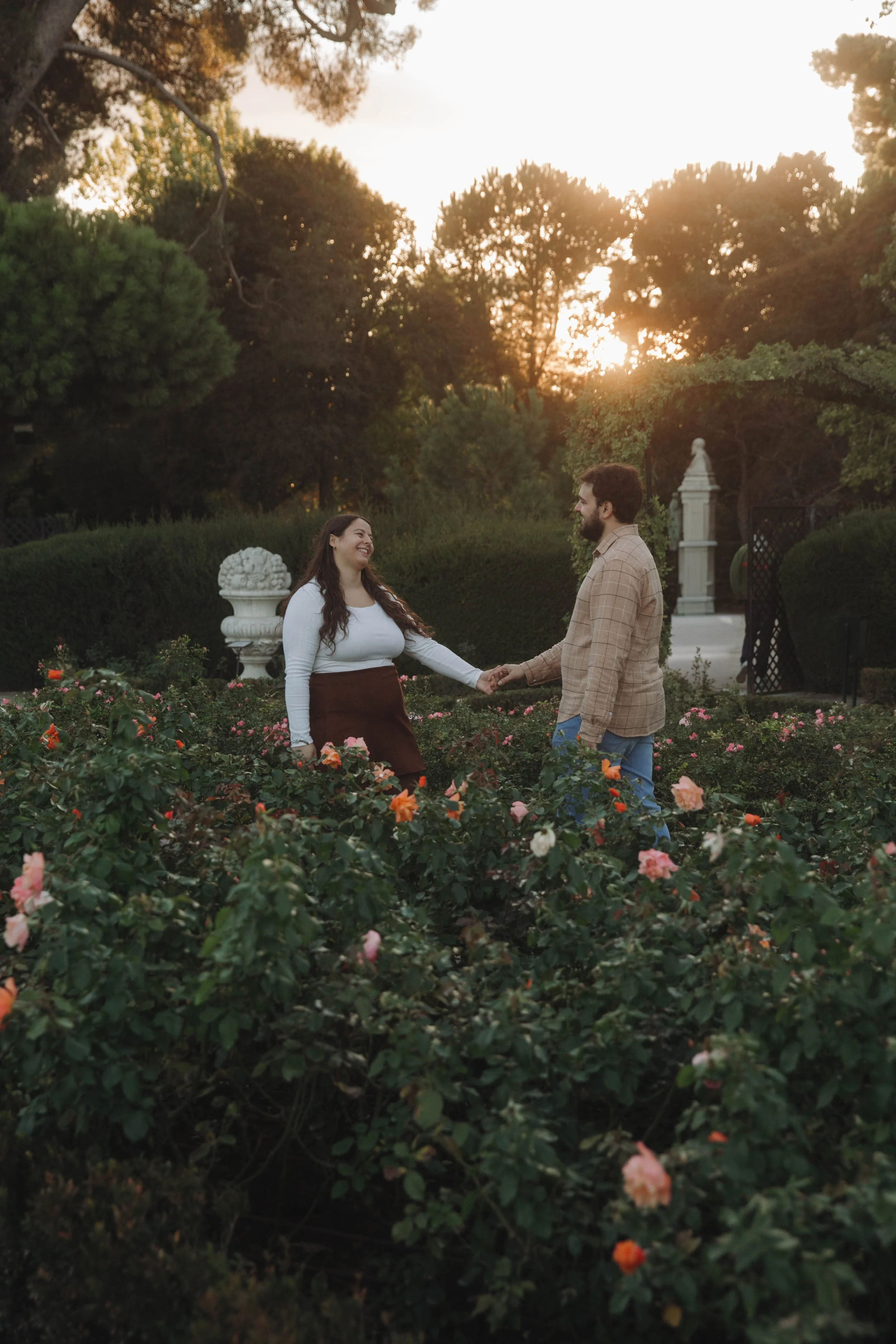 A couple holding hands and smiling in a garden at sunset, surrounded by blooming roses and trees.