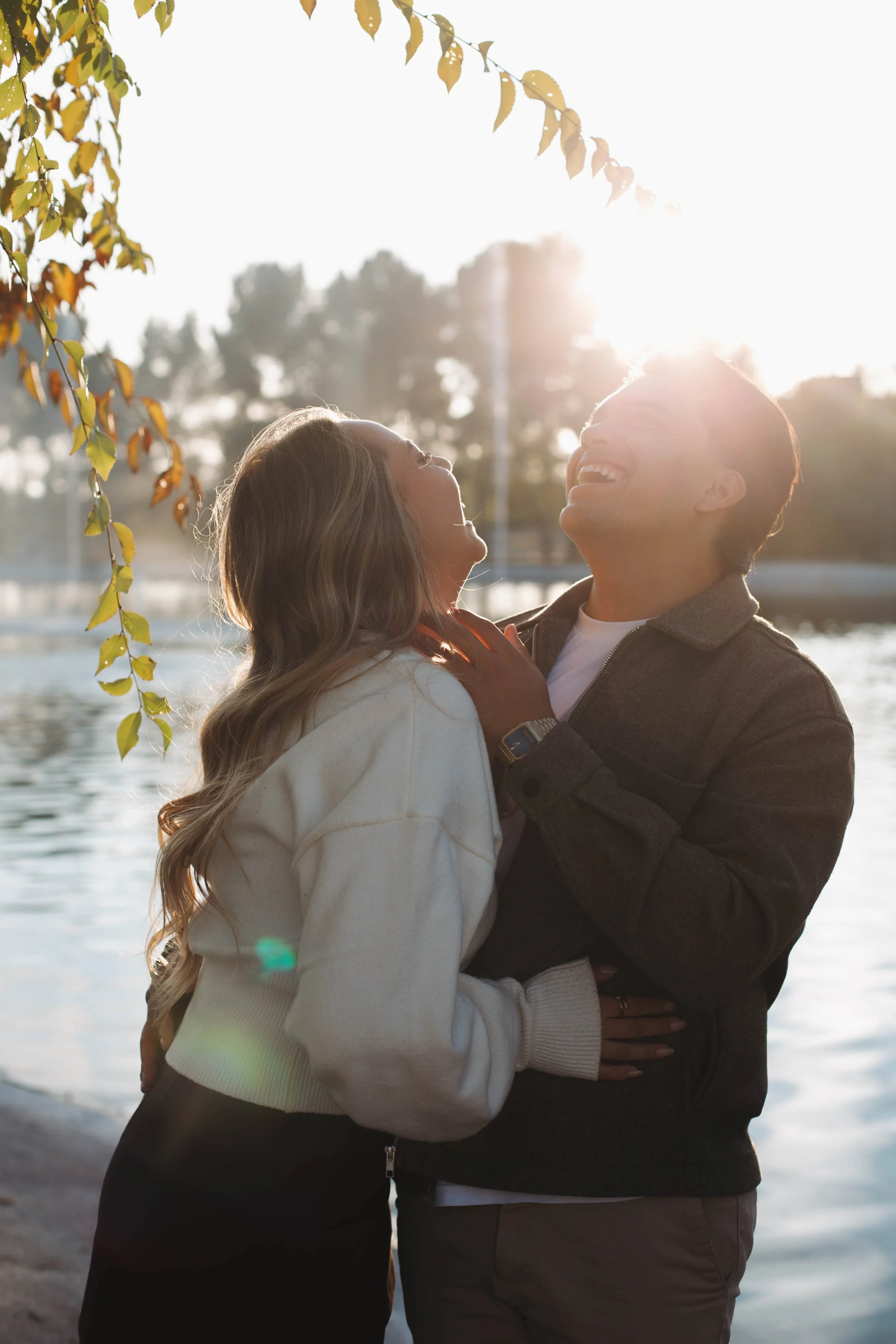A couple laughing and embracing near a body of water, with sunlight behind them and autumn leaves in the foreground in Madrid.