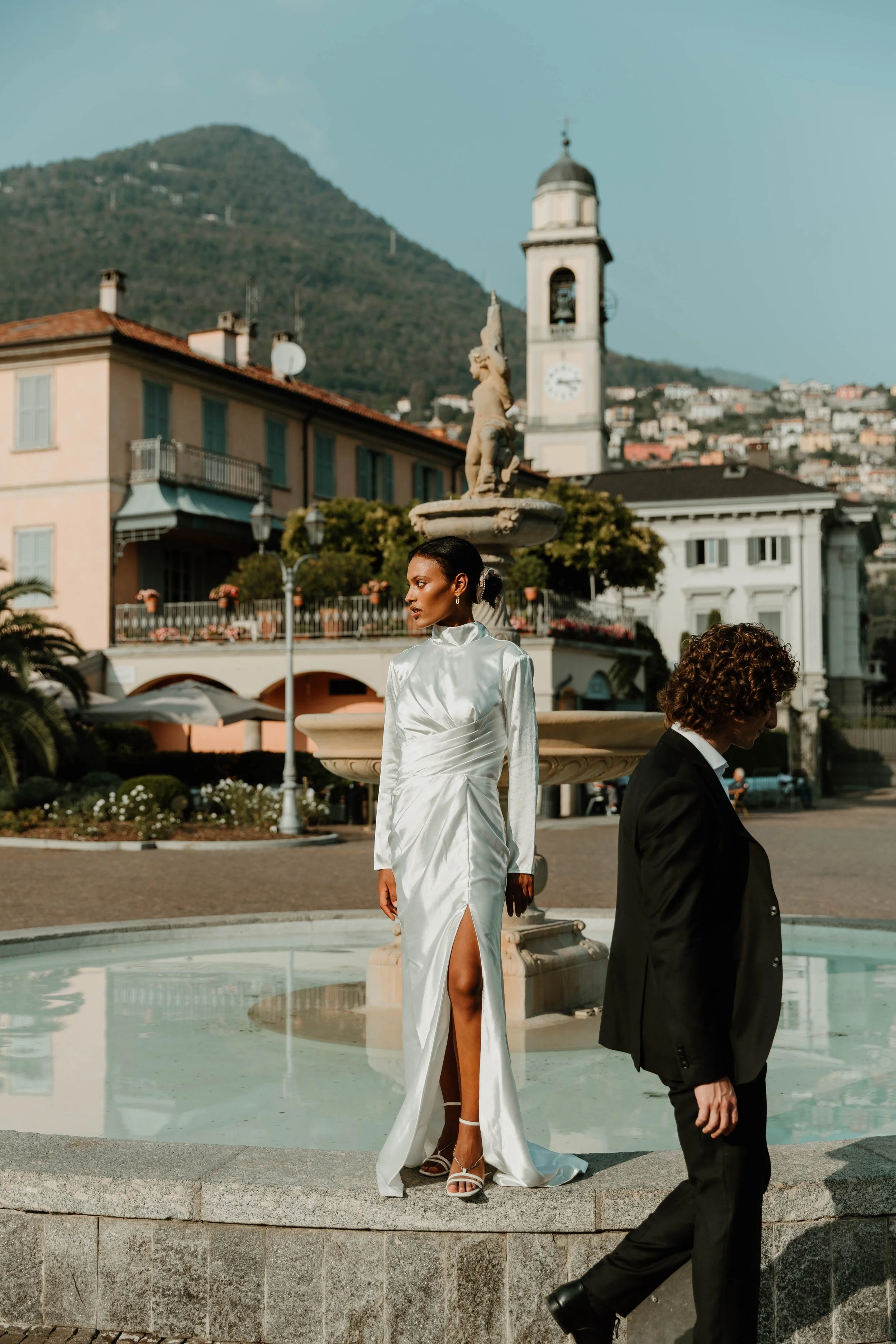 Bride in a silk gown standing on the fountain edge in Cernobbio with pastel buildings behind