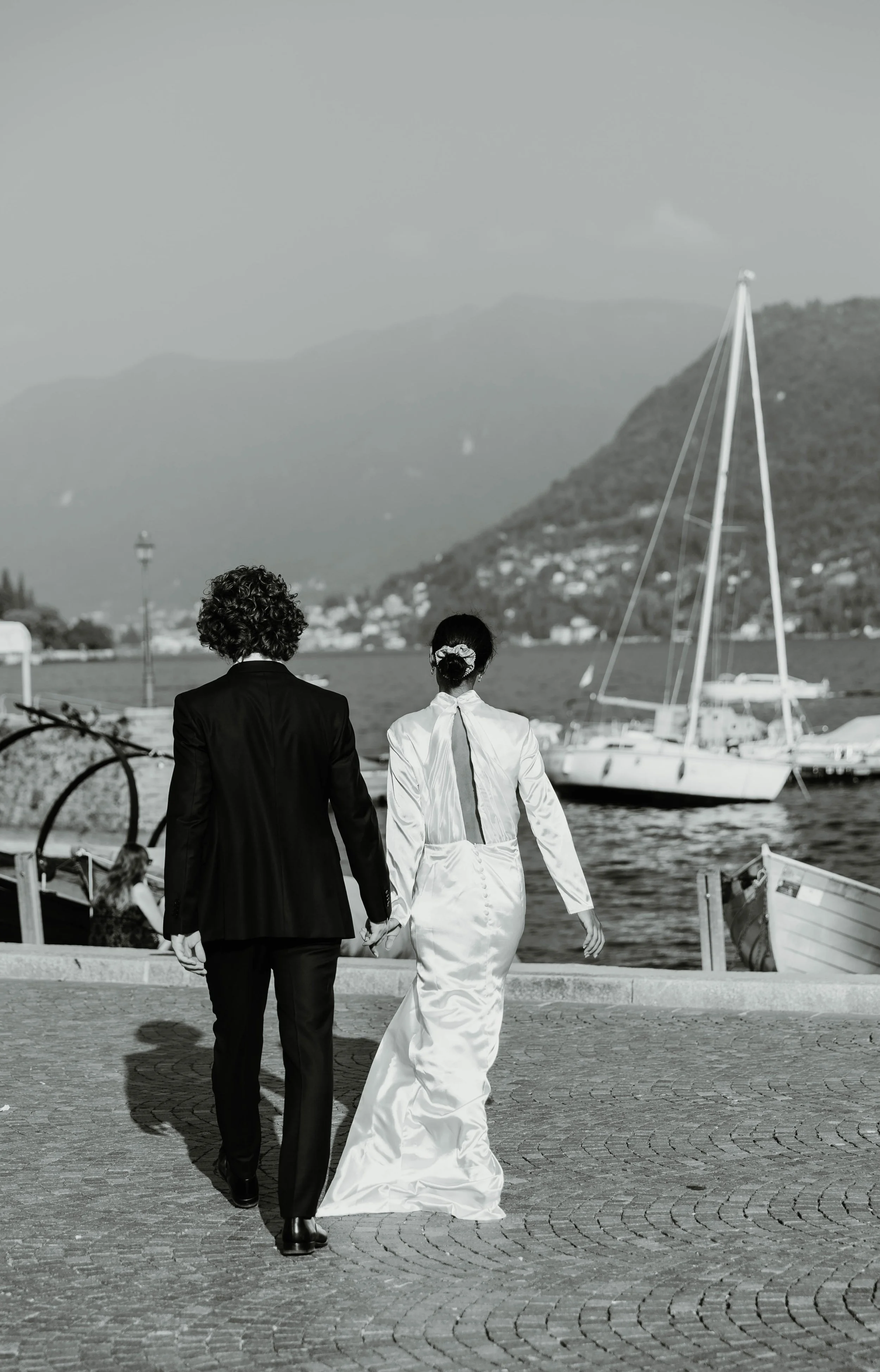 Black and white photo of a couple walking hand in hand toward the boats on Lake Como