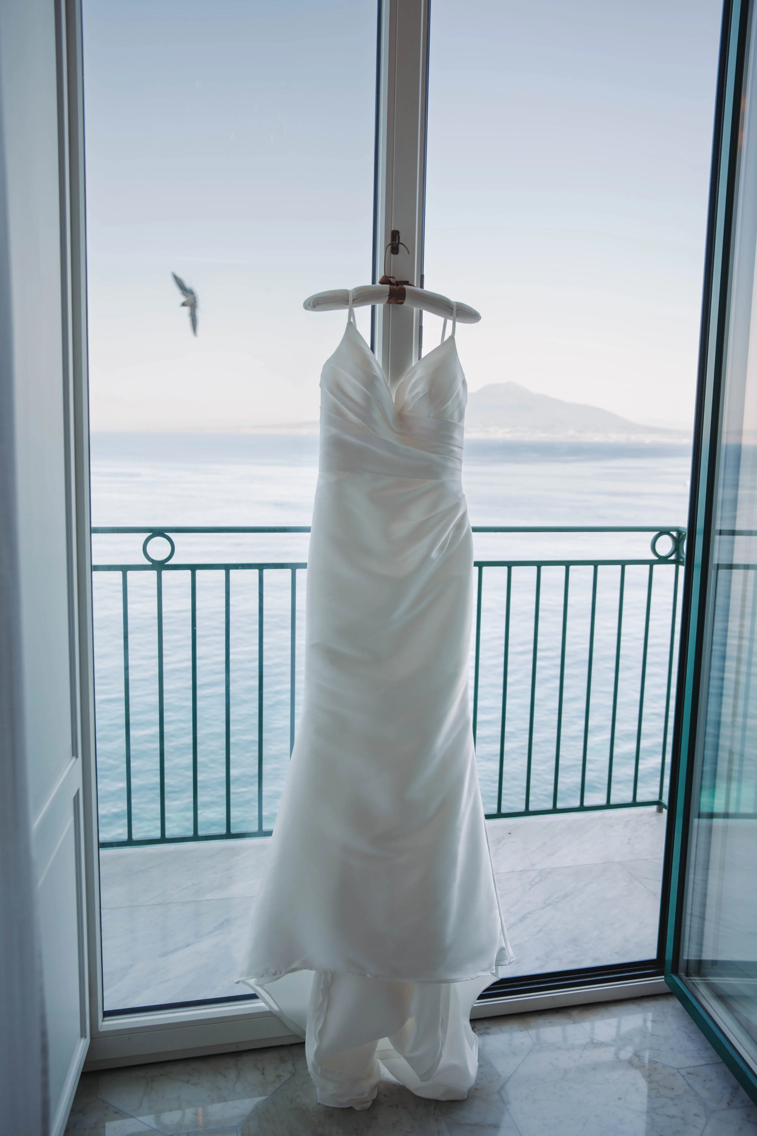 Wedding dress hanging in the balcony doorway with the Bay of Naples behind it