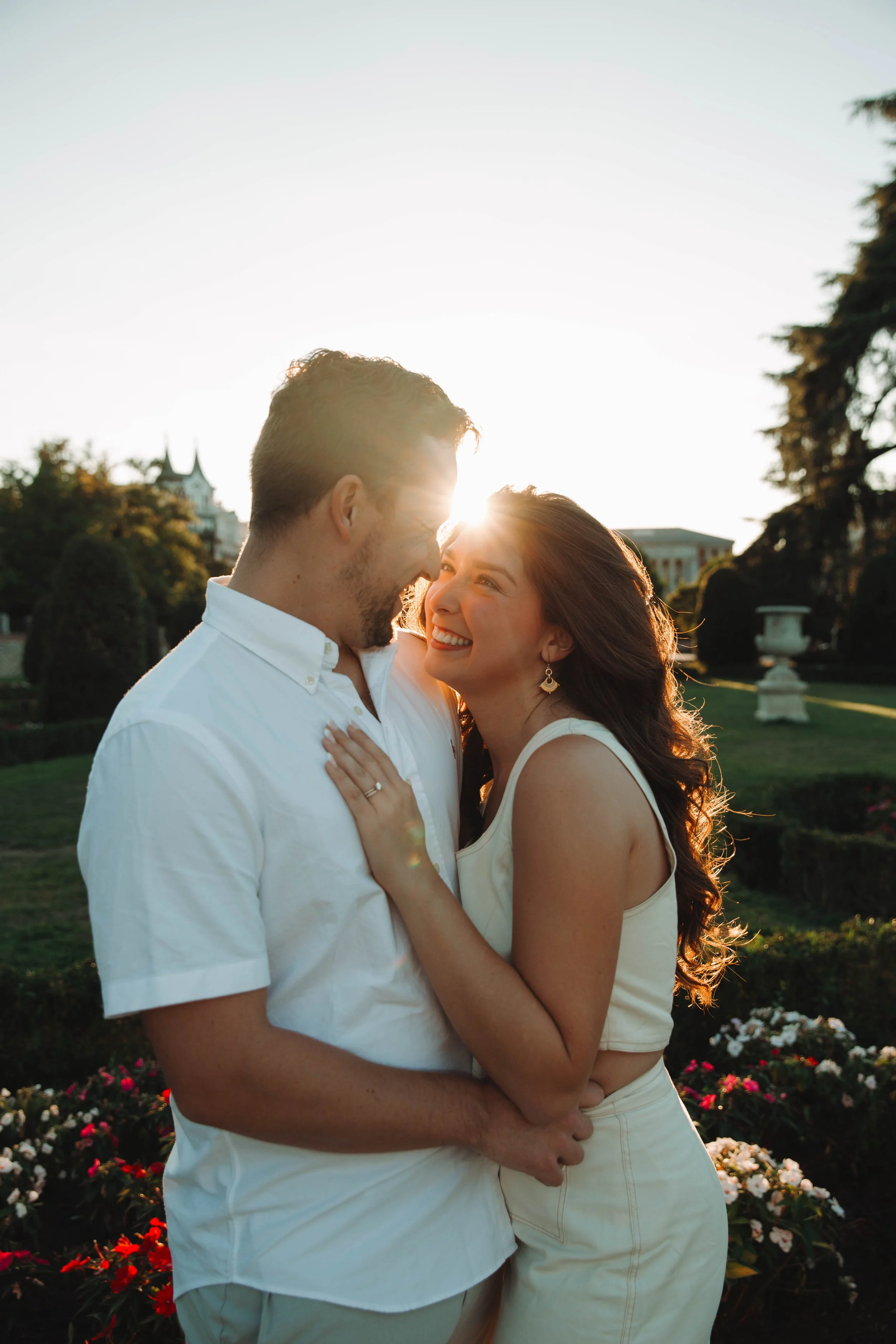 A smiling couple, a man and a woman, is embracing outdoors during sunset with sunburst behind them. in Retiro Park, Madrid They are surrounded by greenery and flowers, dressed in light-colored summer clothes.