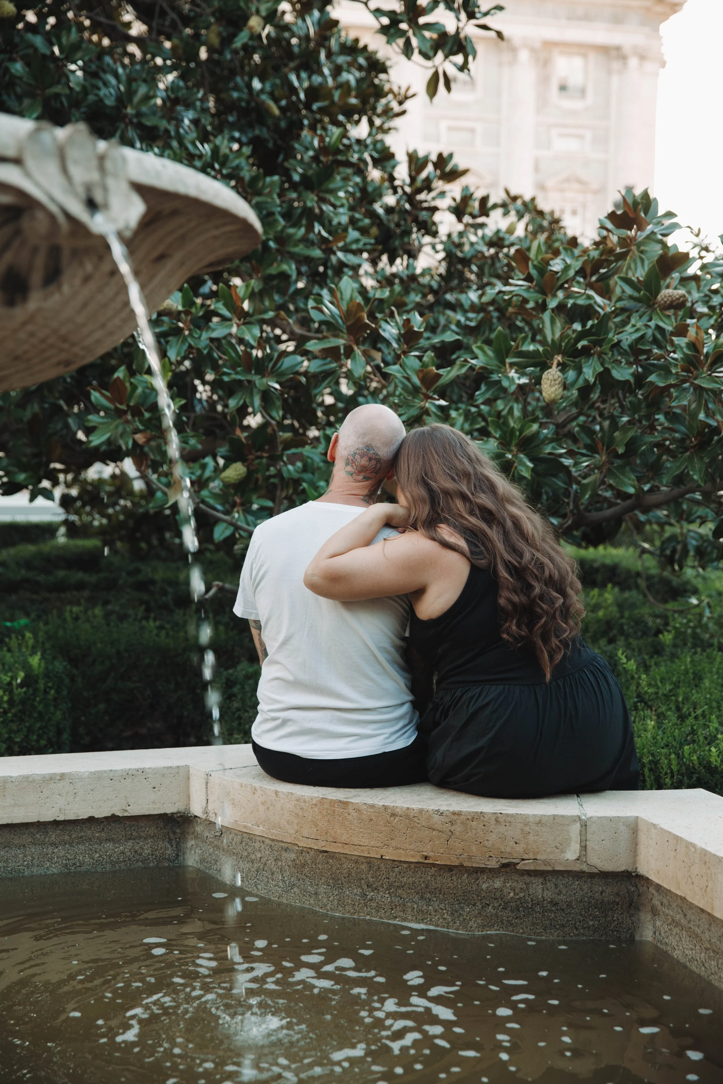 A man and woman sitting close together on a stone ledge near a fountain with water flowing from a bird bath. The man is bald with tattoos on his neck and wearing a white t-shirt, while the woman has long wavy brown hair and is wearing a black dress. They are embracing and leaning into each other as they sit outdoors near green bushes and trees.