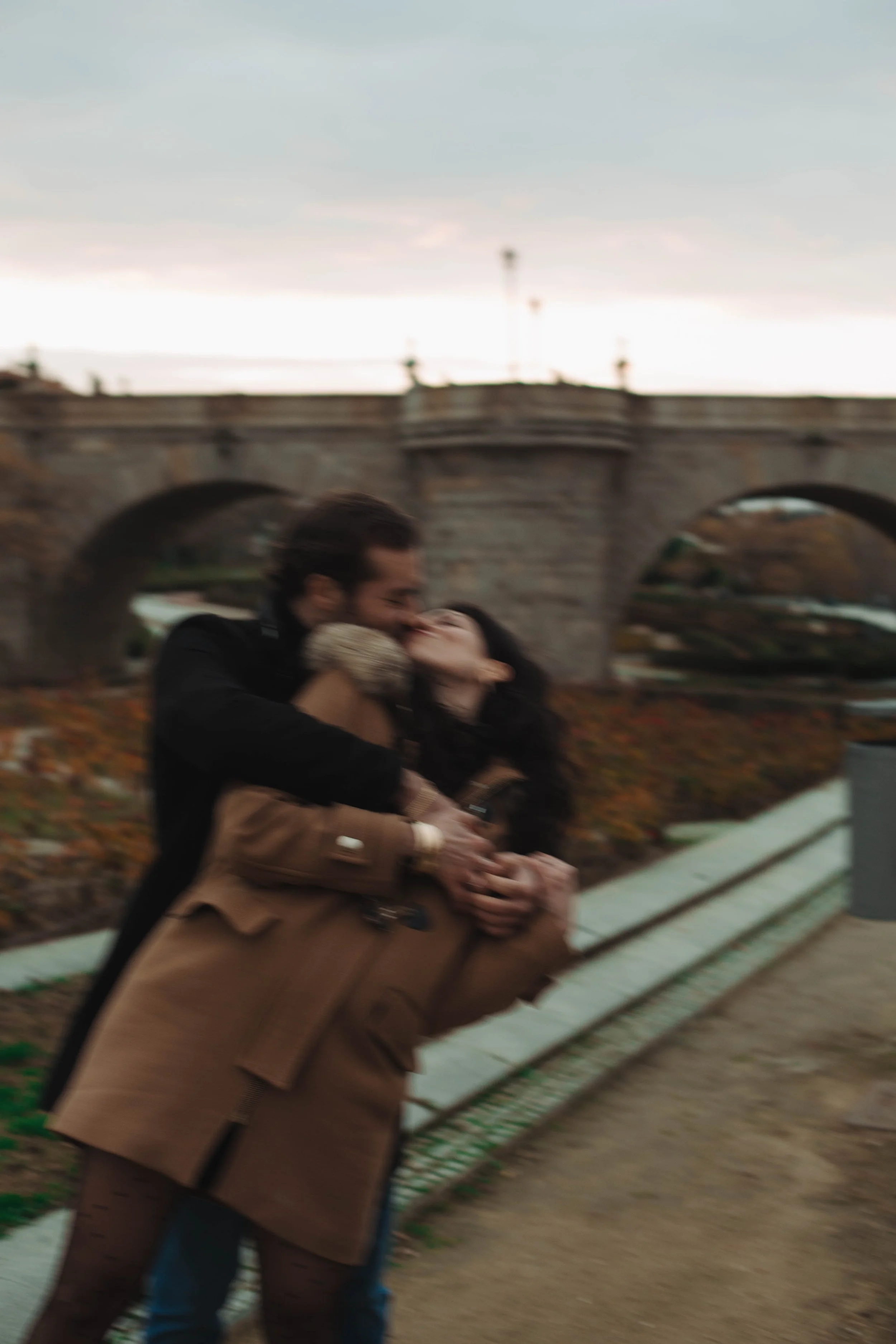 A blurry photo of a couple embracing and kissing outdoors, with a stone bridge in the background during evening or late afternoon in Madrid.