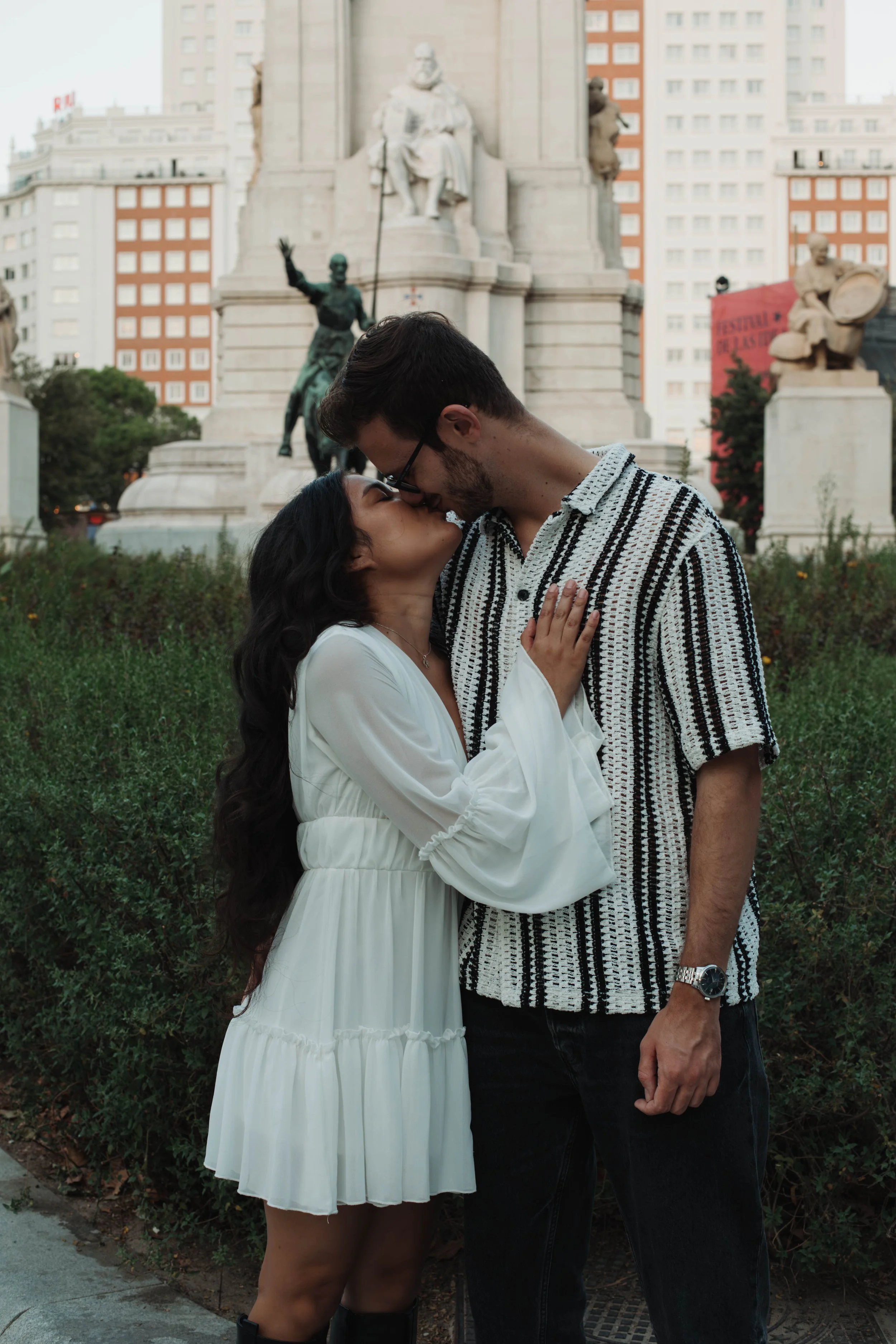A couple sharing a kiss in front of a monument with sculptures and buildings in the background in Madrid near the Royal Palace.