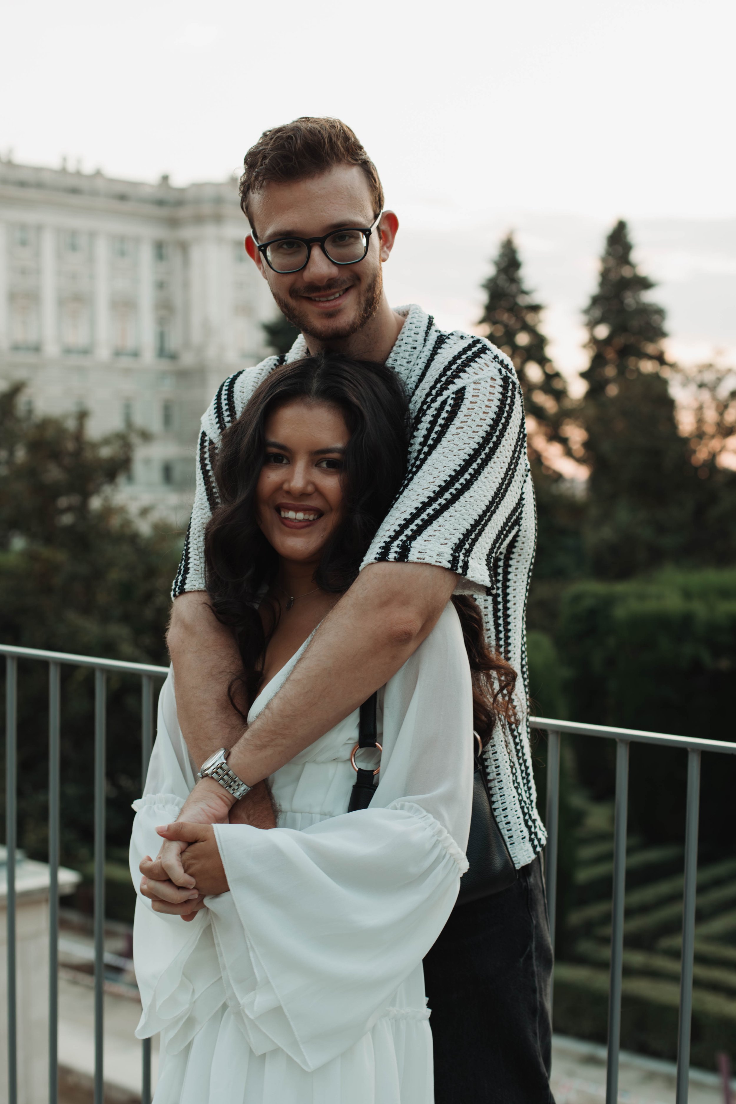 A young couple standing outdoors on a balcony, smiling with the man hugging the woman from behind. The man is wearing glasses and a striped shirt, and the woman is wearing a white dress with long sleeves. Trees and a large building are visible in the background during sunset