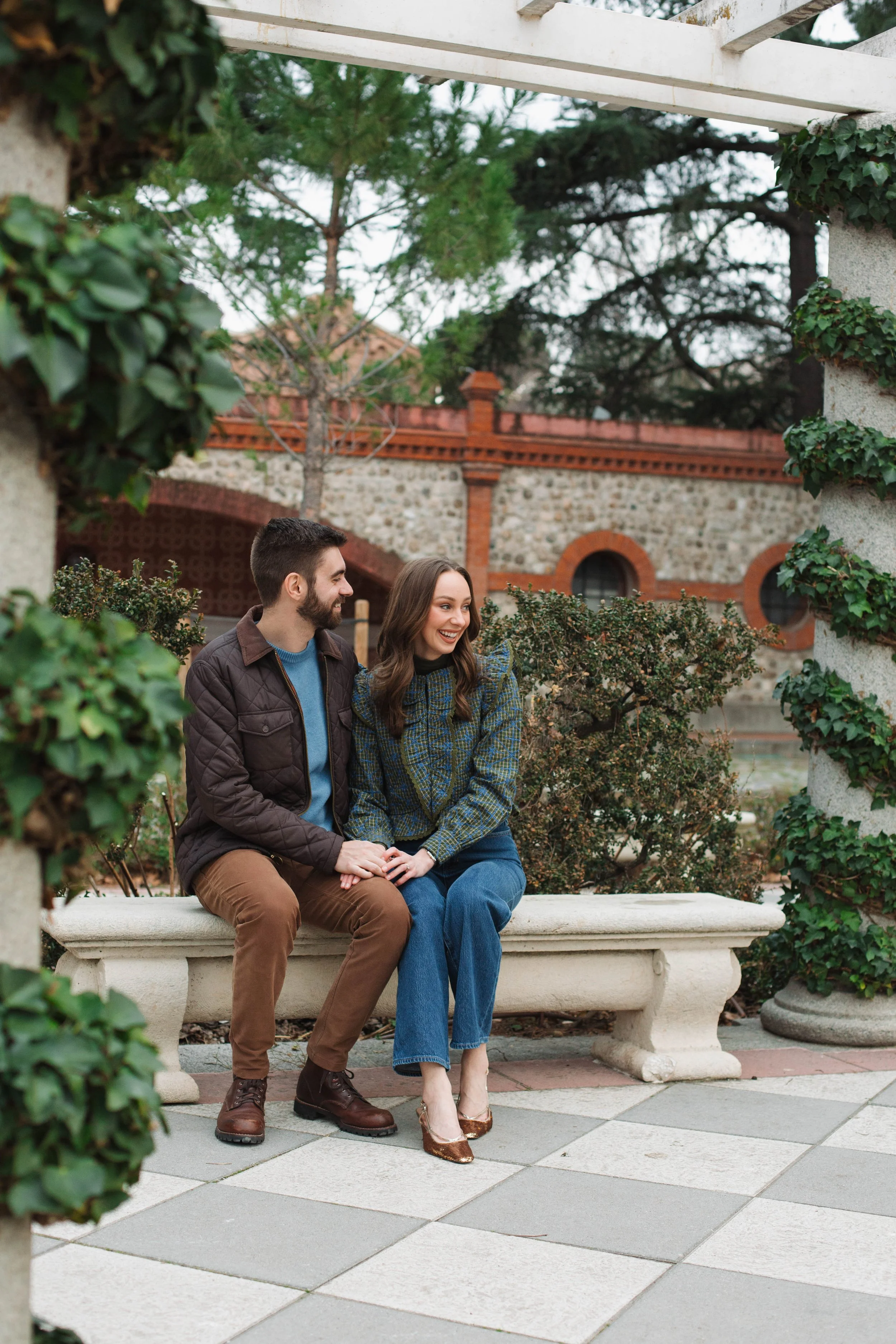 A couple sitting on a stone bench in an outdoor garden, smiling and holding hands, surrounded by greenery and architectural structures.