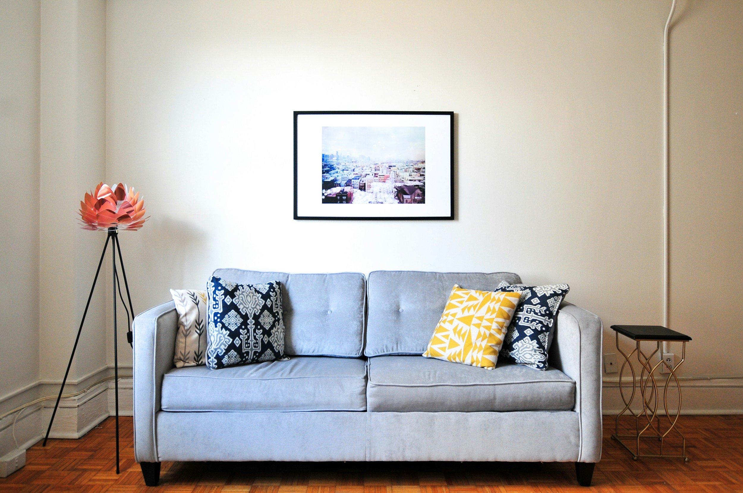 Modern gray sofa with blue and yellow pillows and a pink floor lamp. Framed cityscape above.