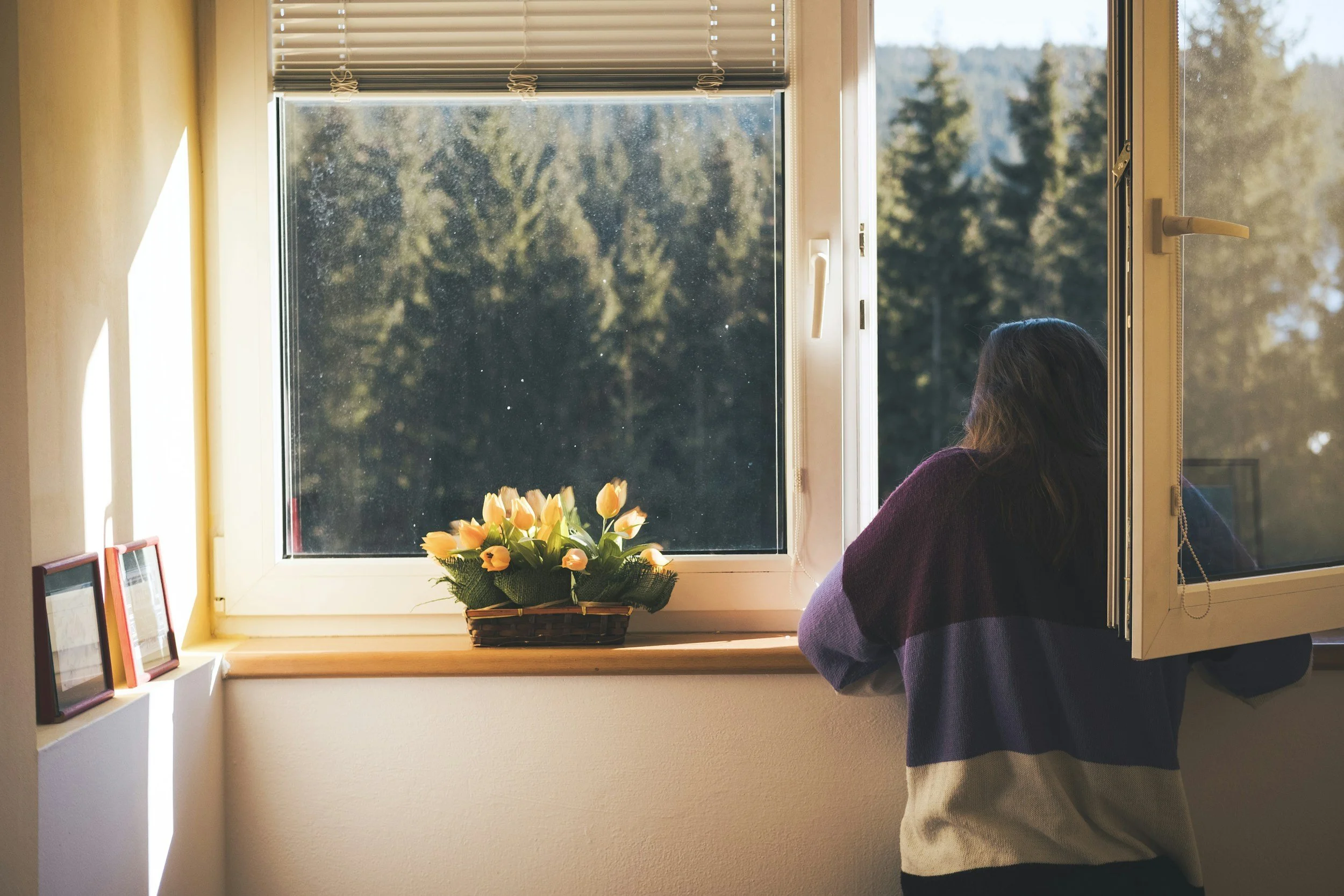 Woman in purple striped shirt looking out window with yellow tulips on window sill after anxiety & people-pleasing therapy