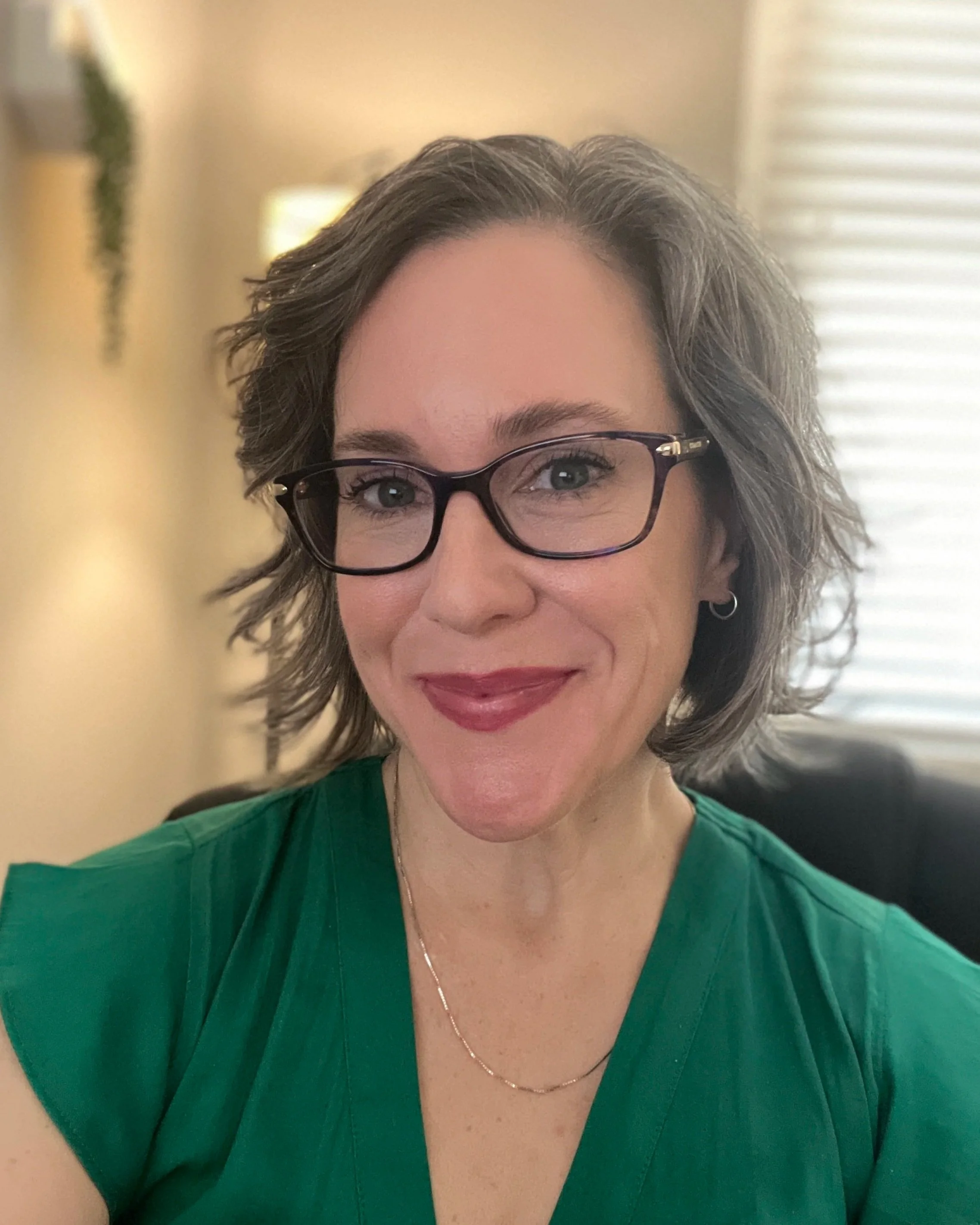 A woman with short, curly brown hair, glasses, and a green top smiling at the camera, sitting indoors with a blurred background including blinds and a plant.
