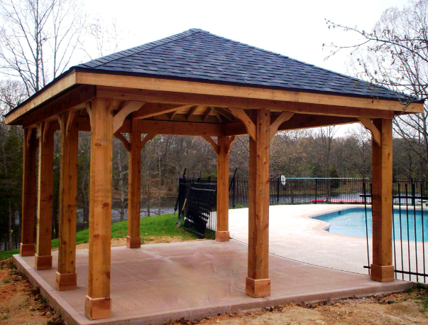 Newly constructed Wooden gazebo near a swimming pool with a concrete deck, black fence, and trees in the background.